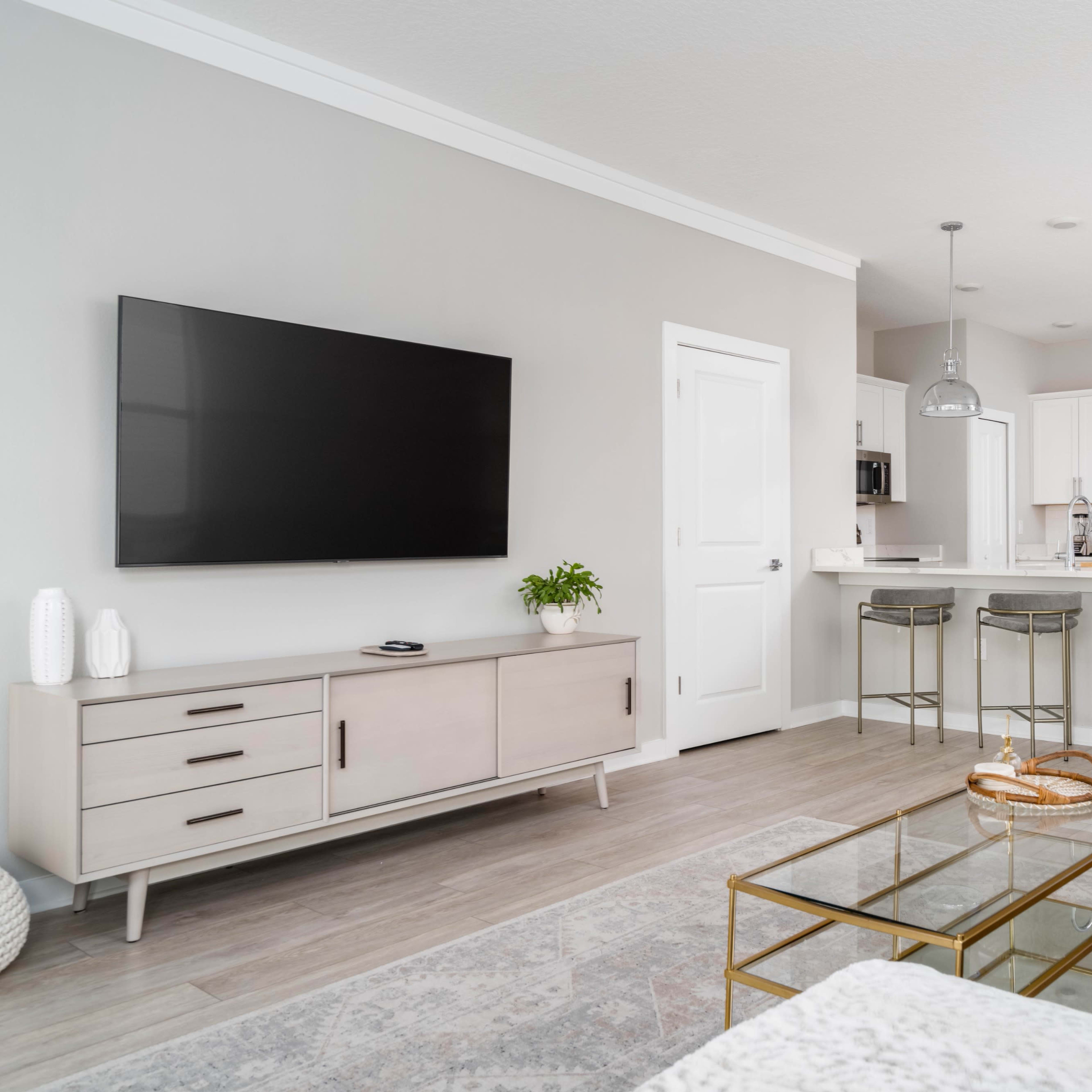 A modern living room features a flat-screen TV mounted on a light-colored wall, with a kitchen area in the background that includes white cabinets and stainless steel appliances.