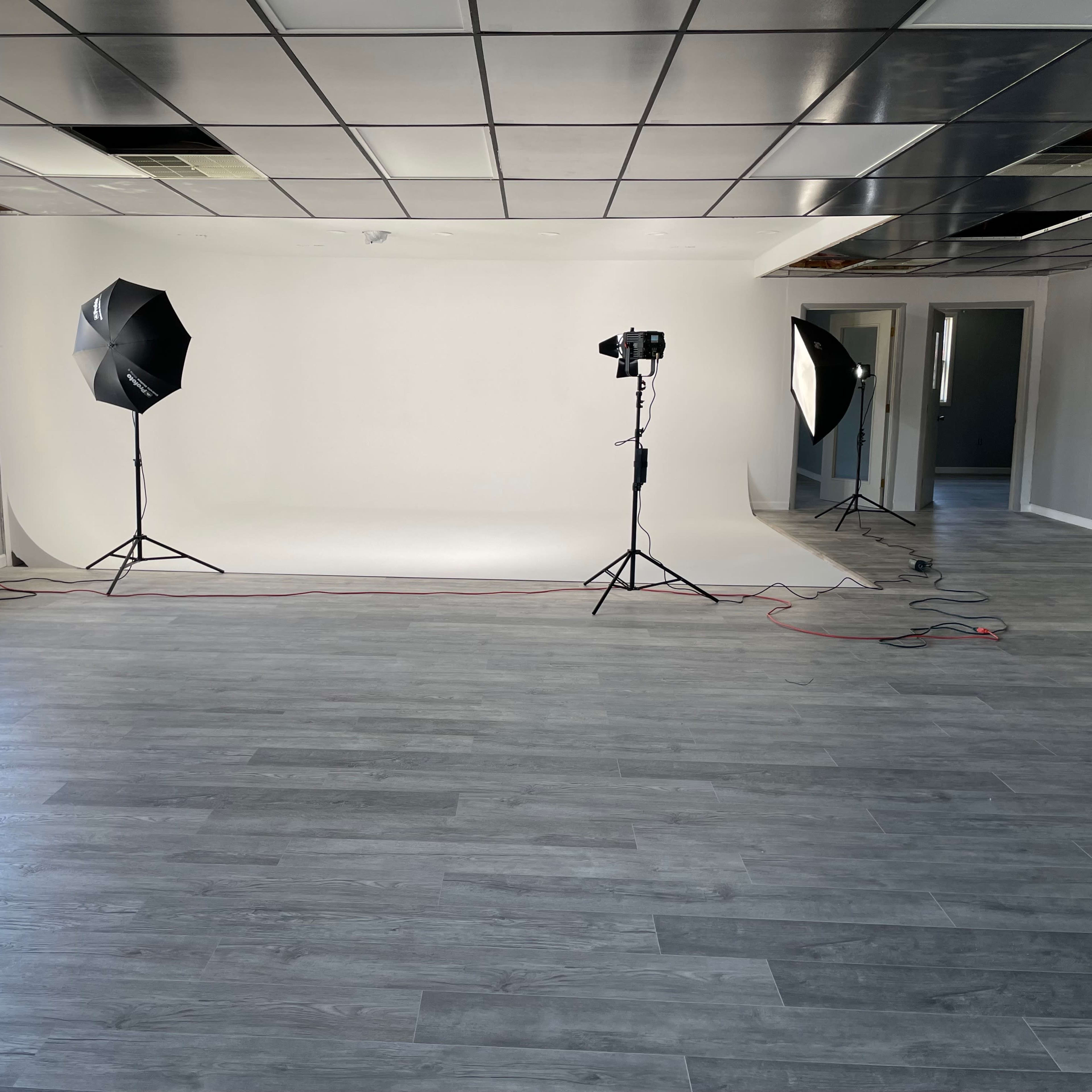 A spacious room with a seamless white backdrop, two studio lights on stands, and gray laminate flooring.