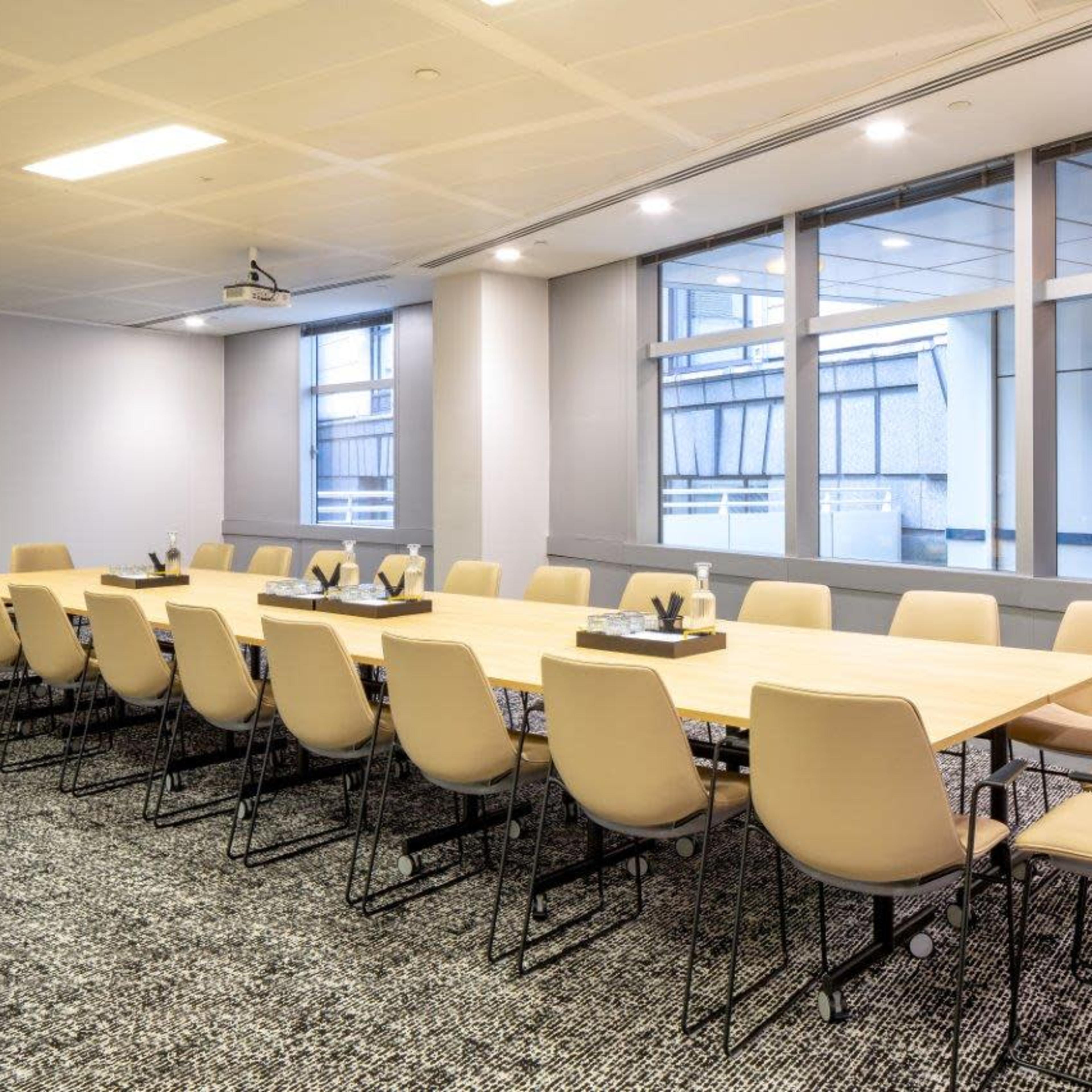 A large conference room features a long wooden table surrounded by beige chairs, with windows providing natural light.