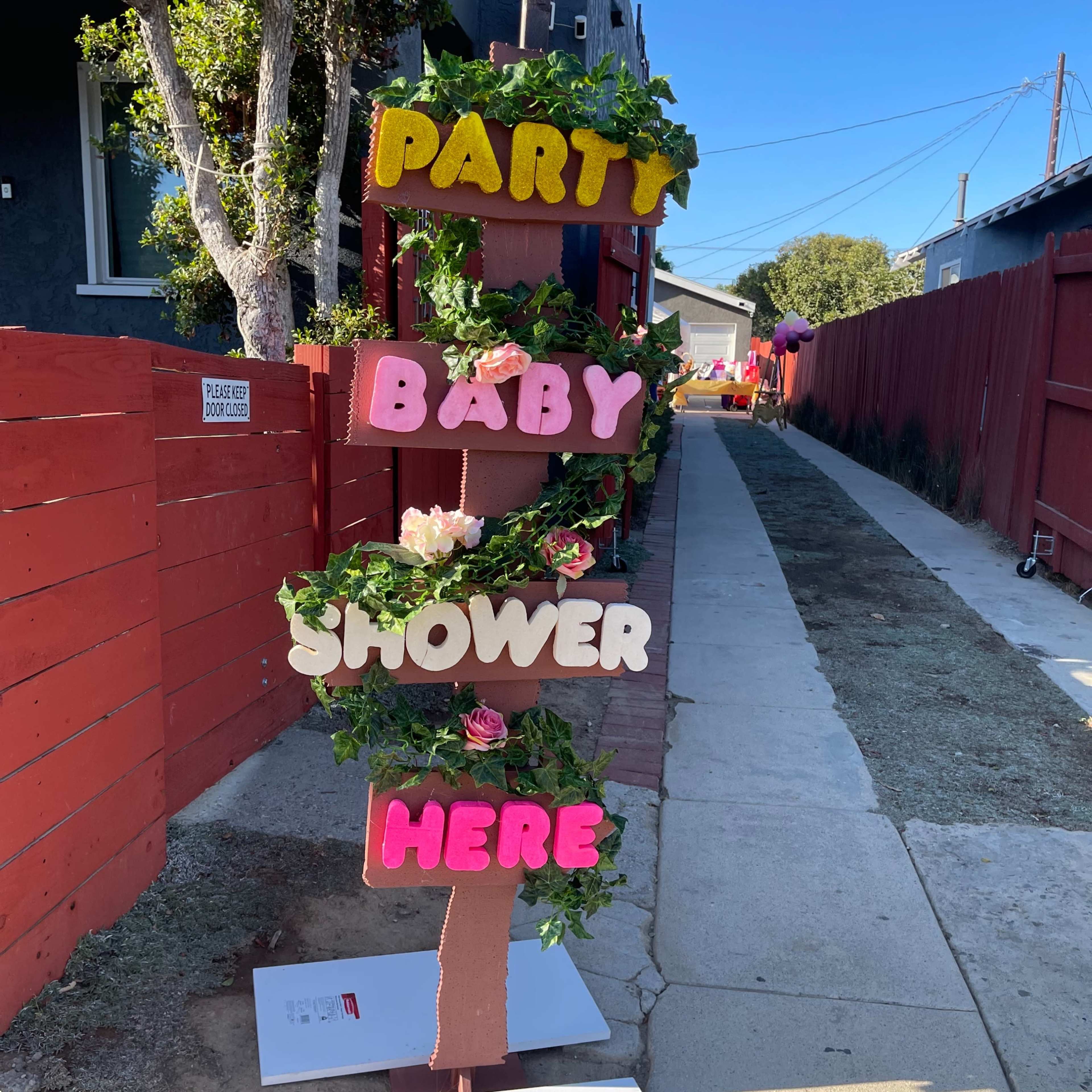 A wooden sign decorated with flowers announces a baby shower, with colorful letters reading "PARTY BABY SHOWER HERE."