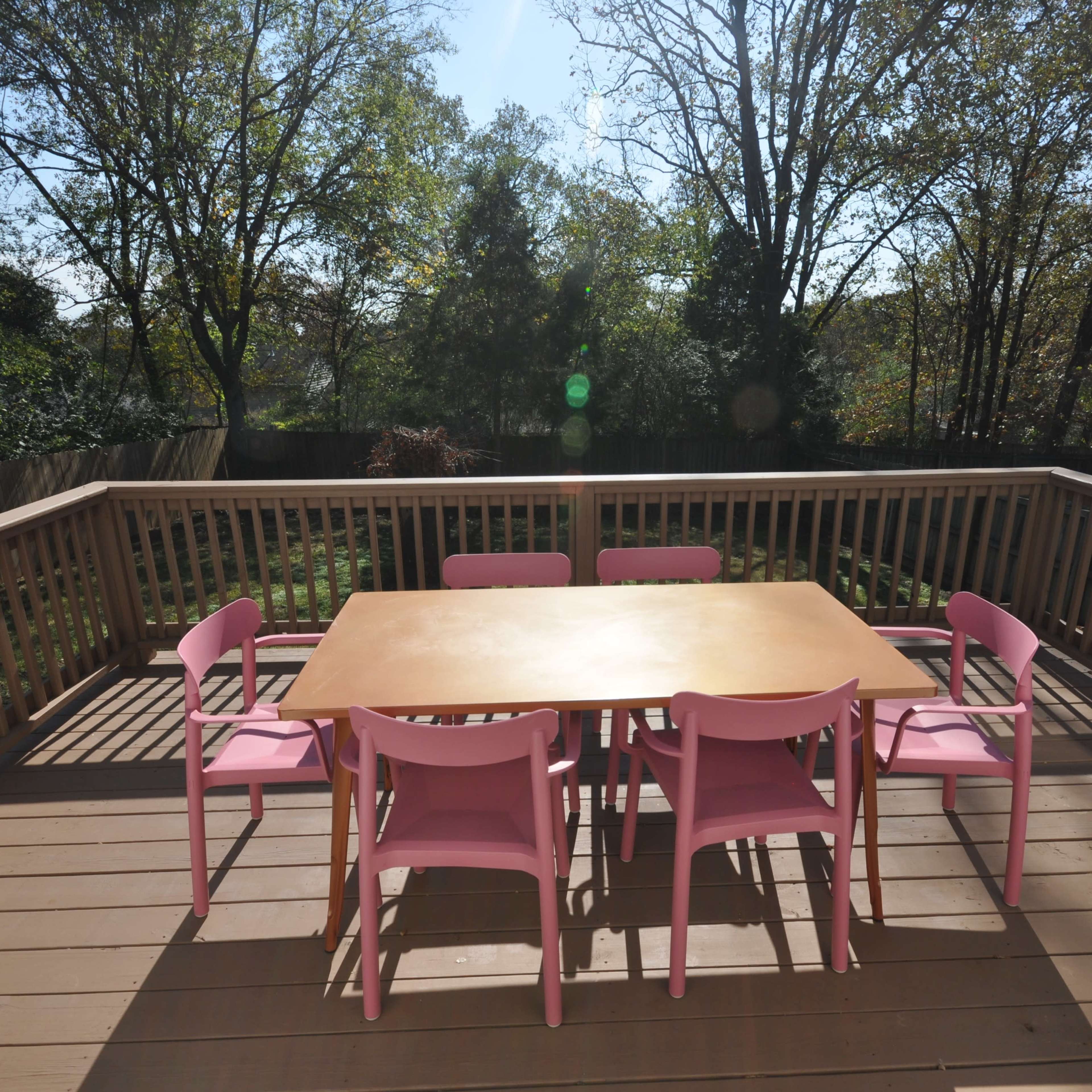 A wooden deck features a large table surrounded by pink chairs, with trees visible in the background.