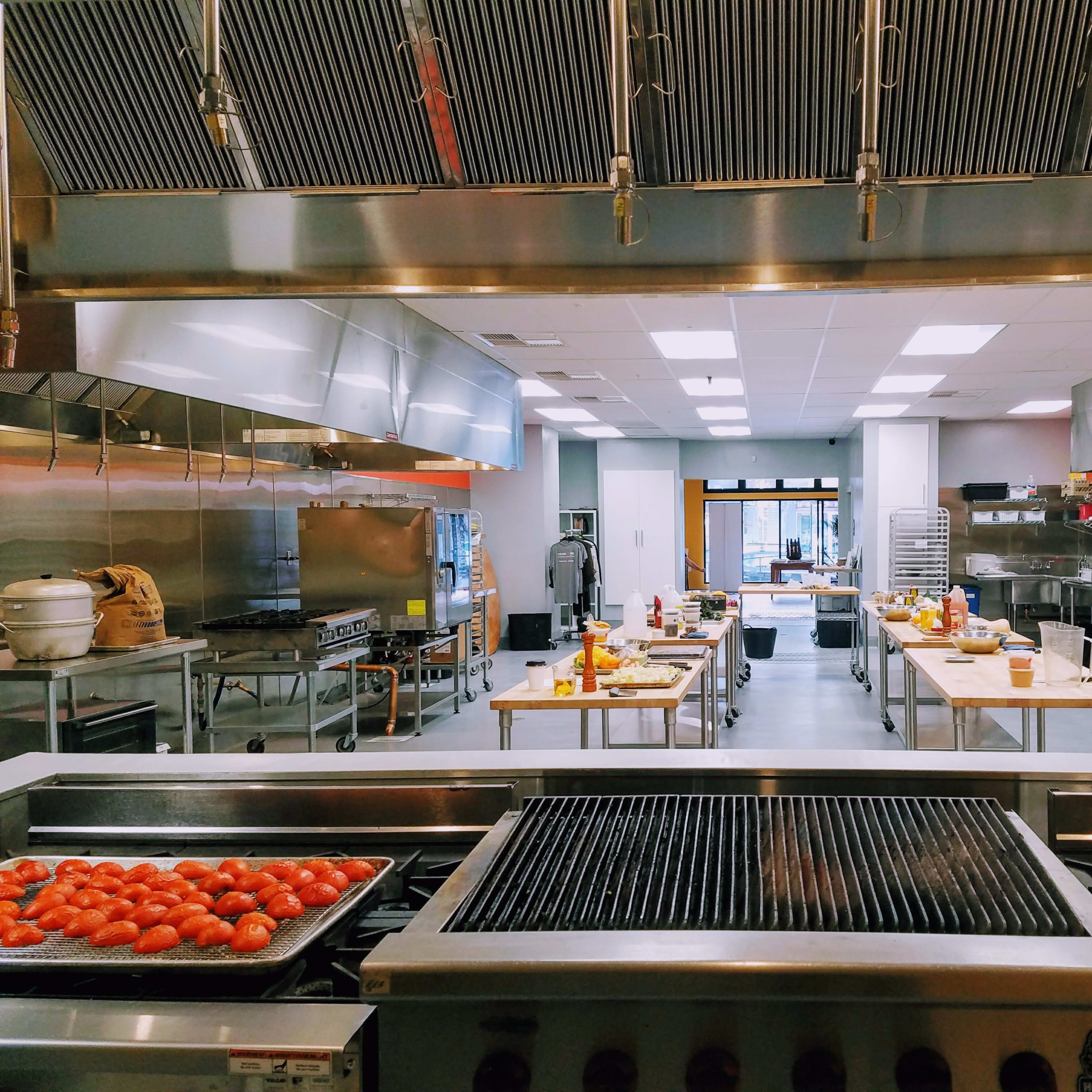 A commercial kitchen is equipped with stainless steel appliances, multiple cooking stations, and large worktables arranged for food preparation.