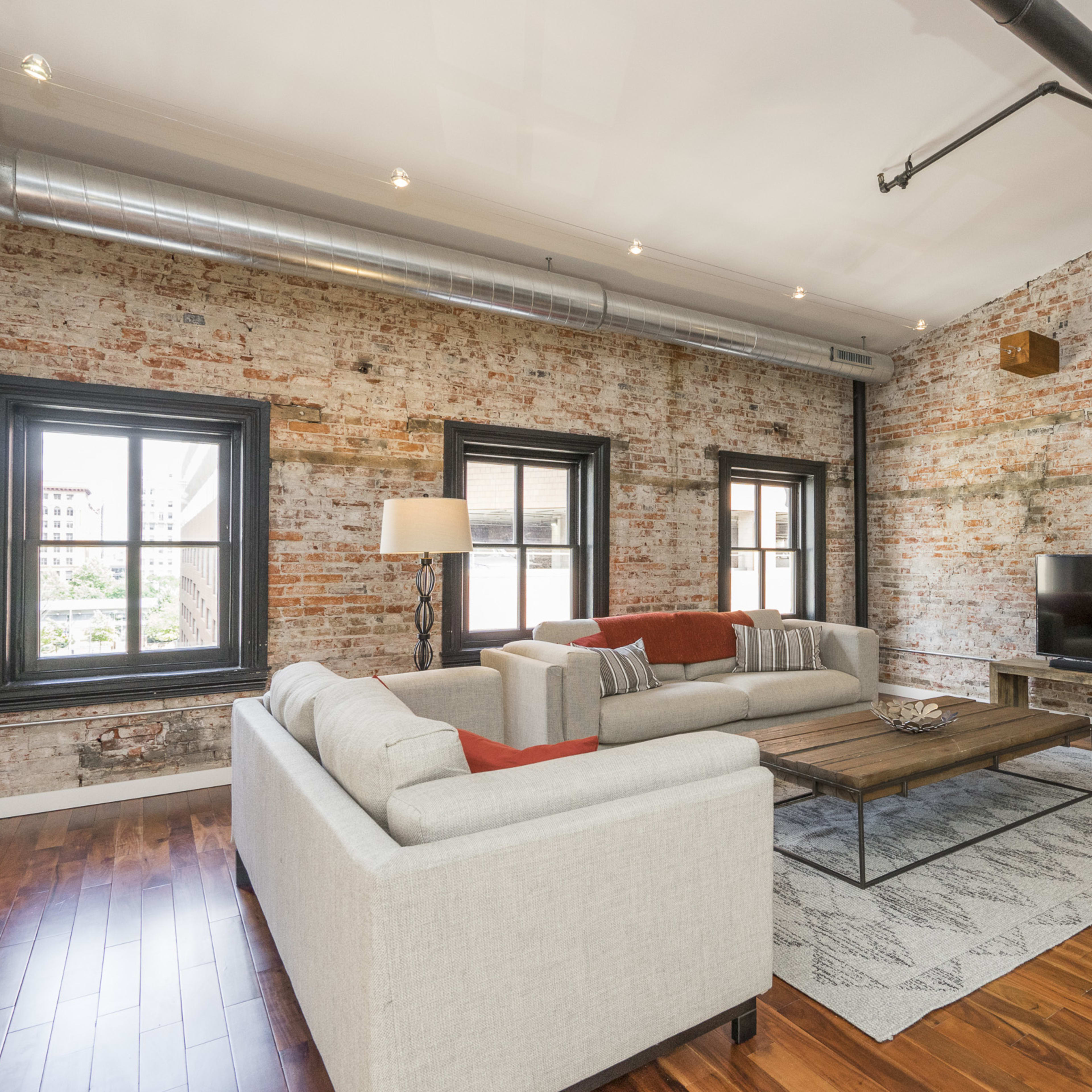 A modern living room with exposed brick walls, hardwood floors, a sectional sofa, a coffee table, and a television mounted on the wall.