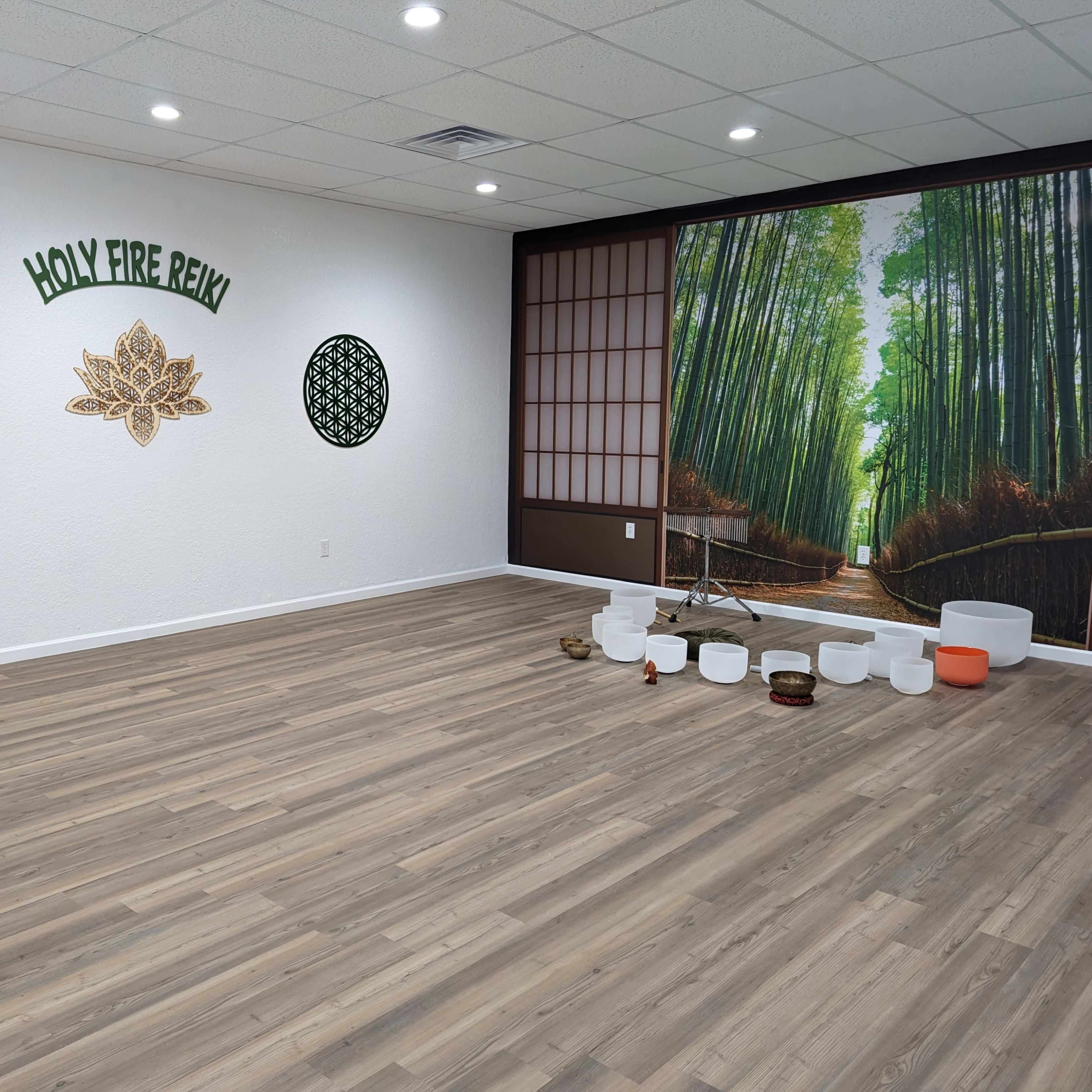 An interior of a meditation or wellness studio featuring wood flooring, a large mural of a bamboo forest, decorative wall elements, and various sound bowls arranged on the floor.