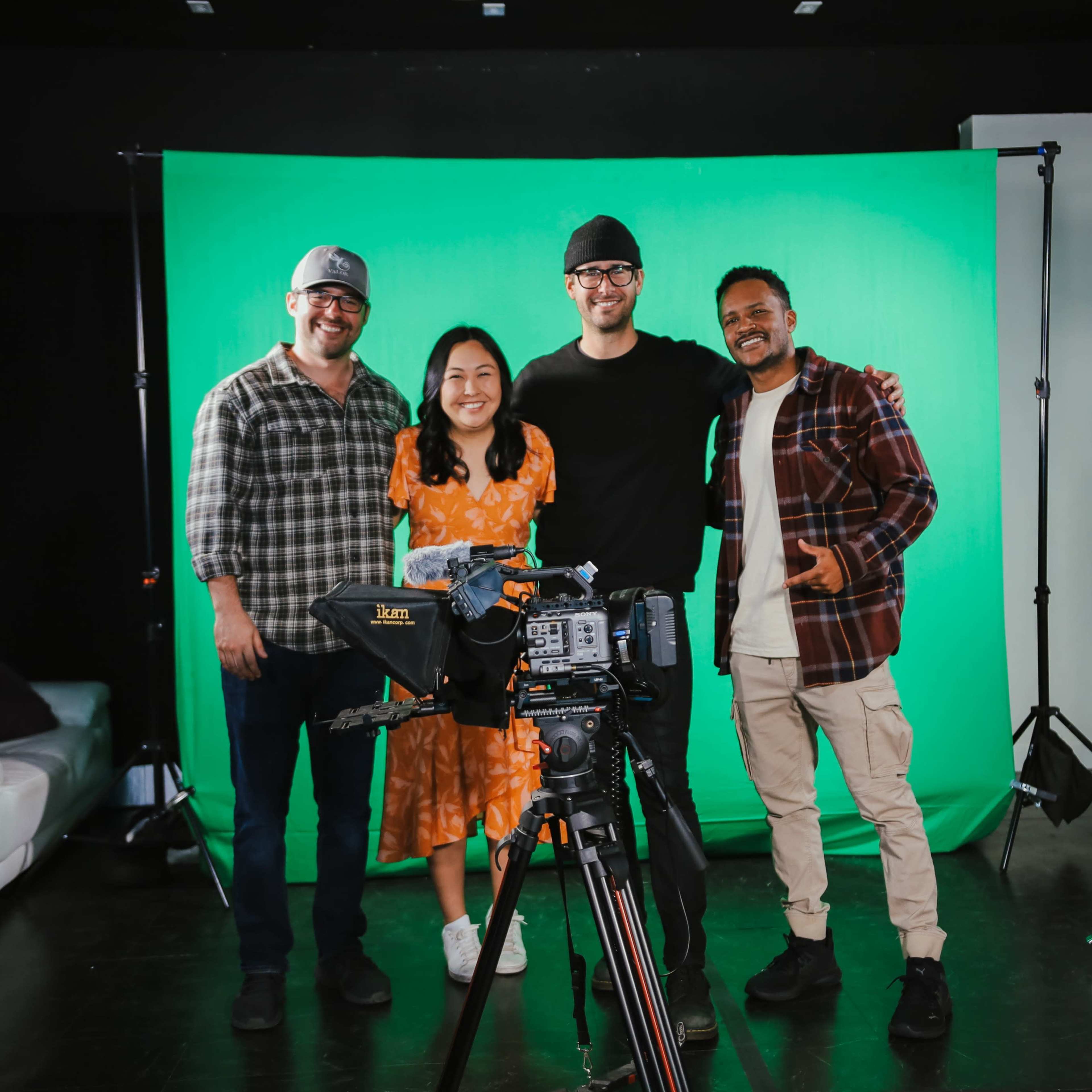 Four individuals pose in front of a green screen, with a camera and lighting equipment set up for a video shoot.