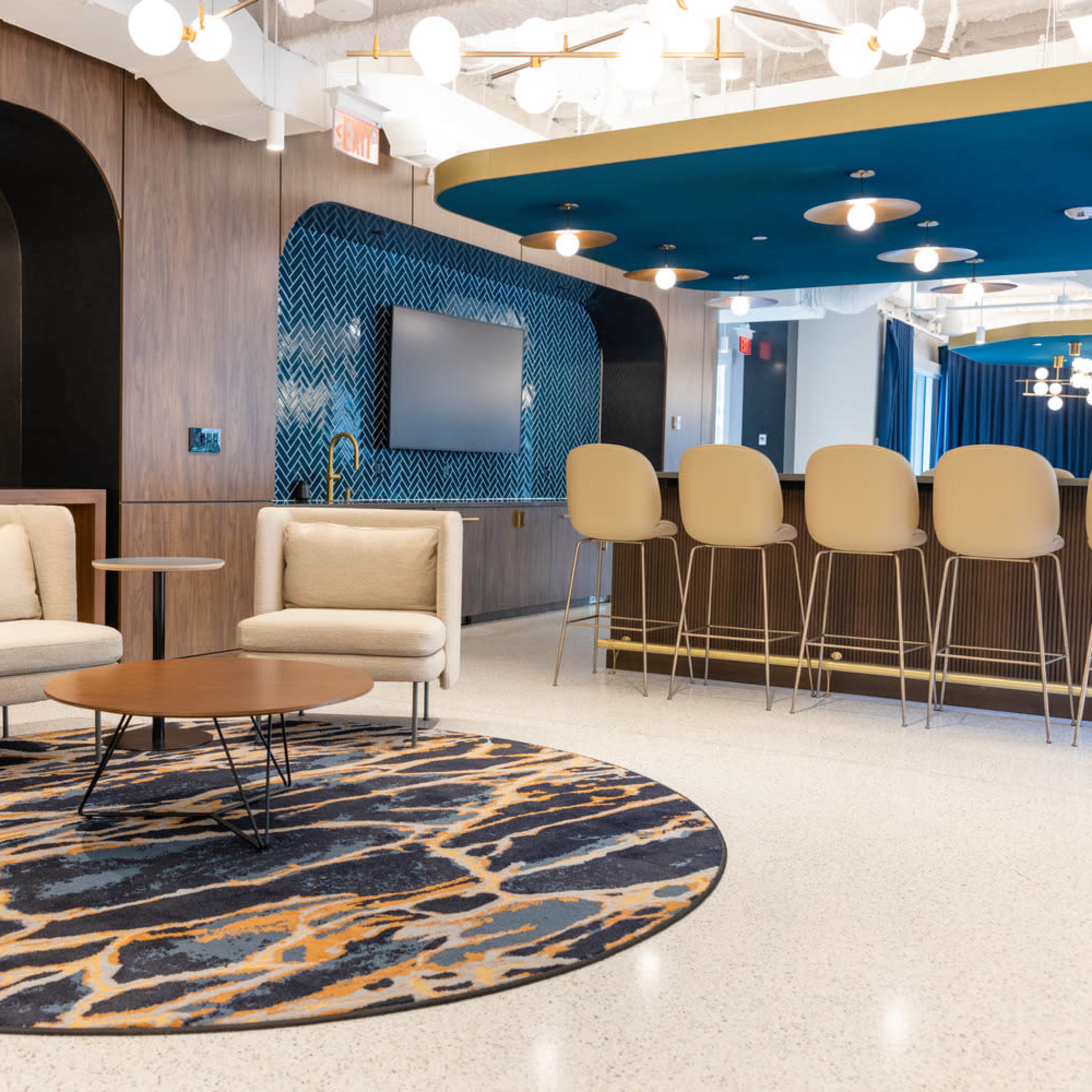 A modern lounge area features light-colored seating arranged around a circular coffee table, with a bar in the background and decorative lighting overhead.