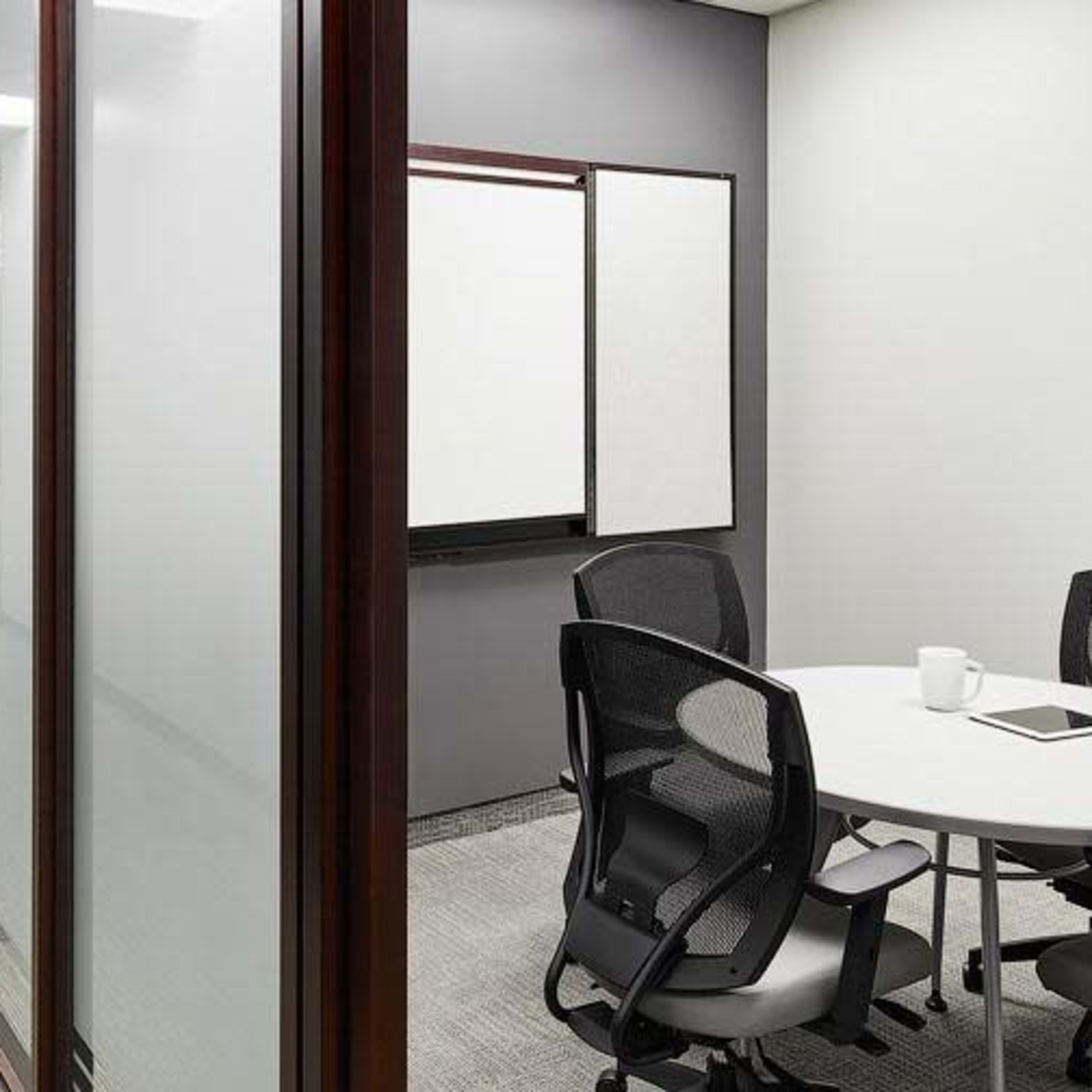 A modern office meeting room with a round table and chairs, flanked by glass walls and a whiteboard.