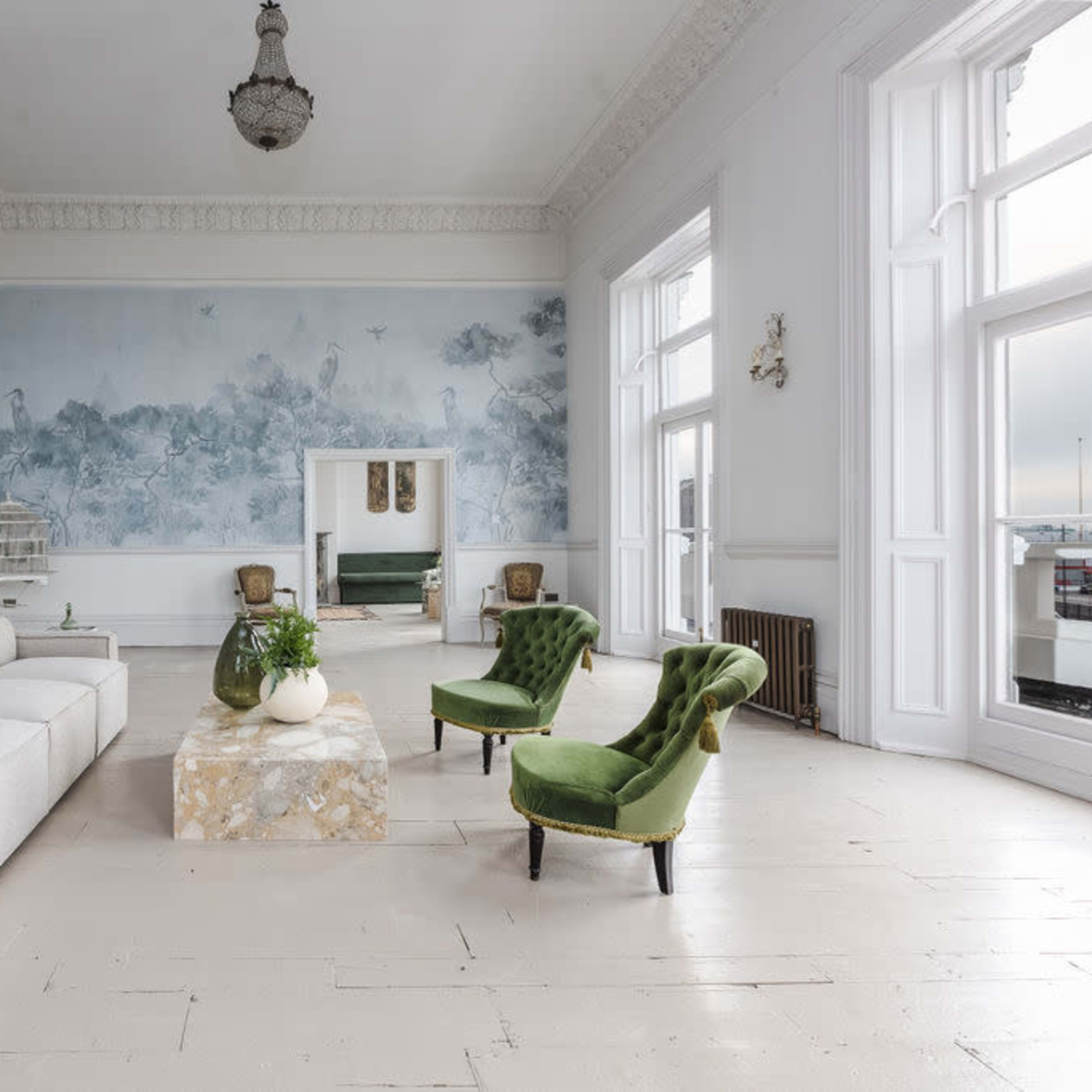 The spacious living room features a light-colored sectional sofa, two green velvet chairs, a marble coffee table, and a large mural depicting a nature scene.