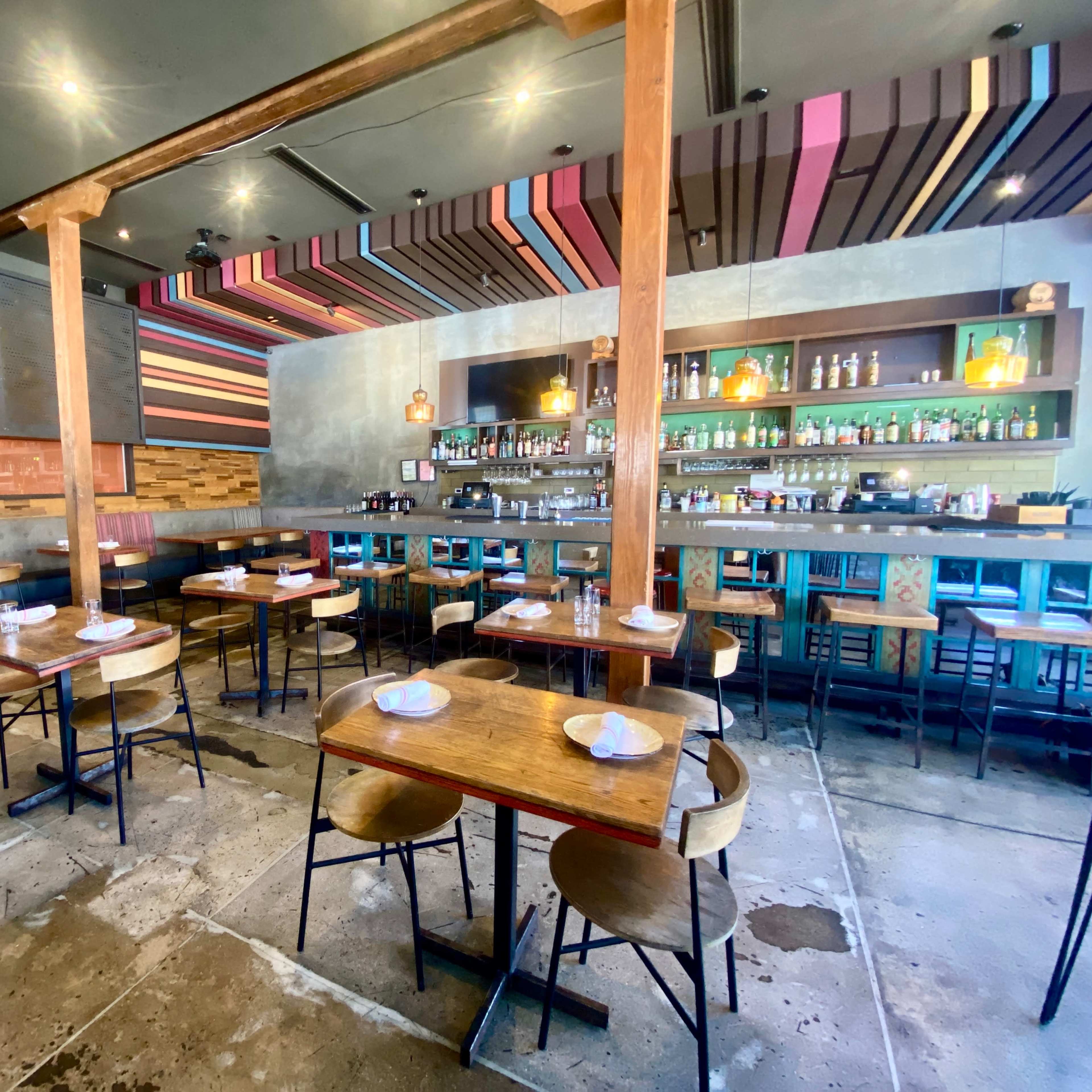 A modern restaurant interior with wooden tables and chairs, a bar area in the back, and colorful striped overhead decorations.