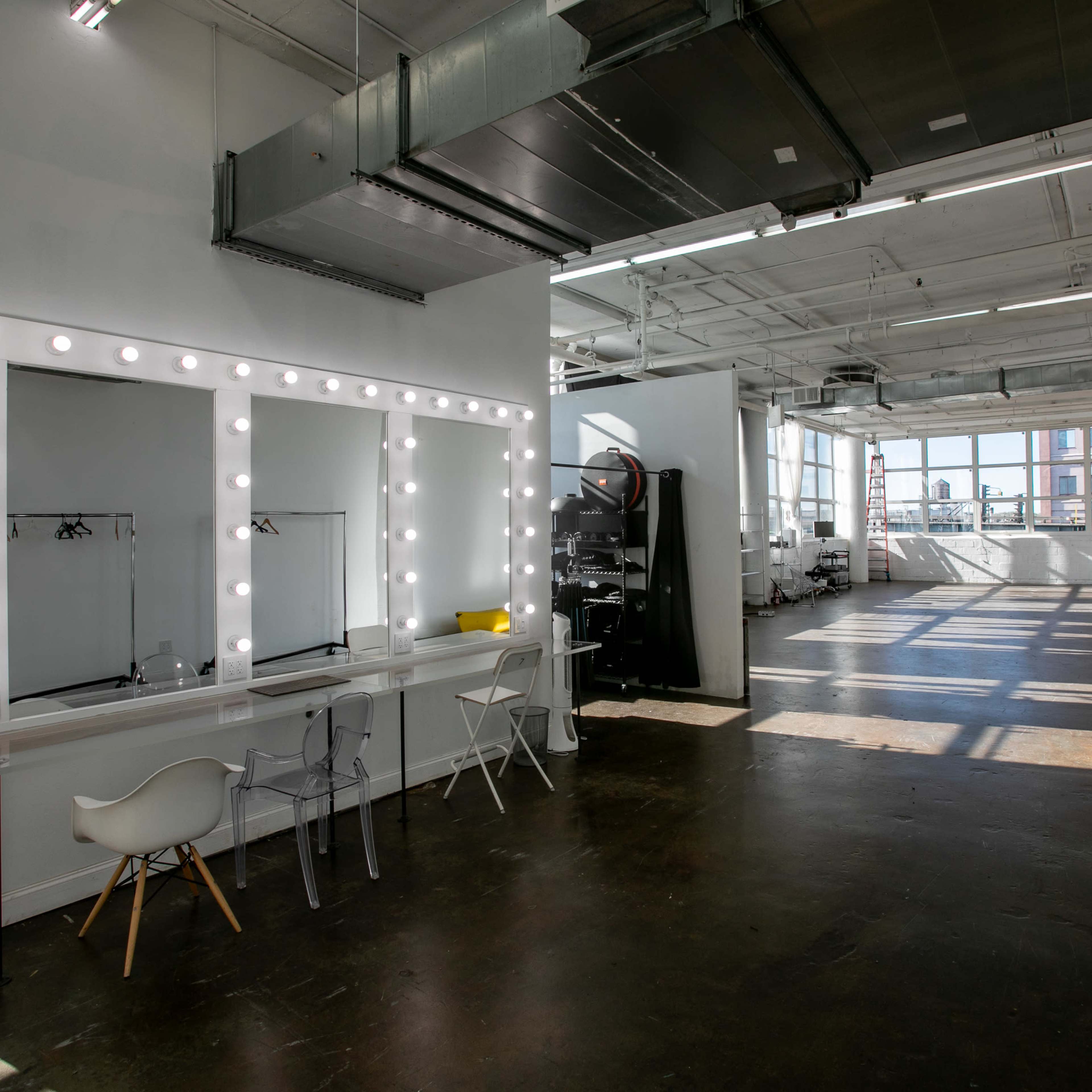 A spacious bright room featuring a large vanity mirror with lights, a white table with chairs, and various furniture pieces against the walls, all illuminated by natural light from wide windows.