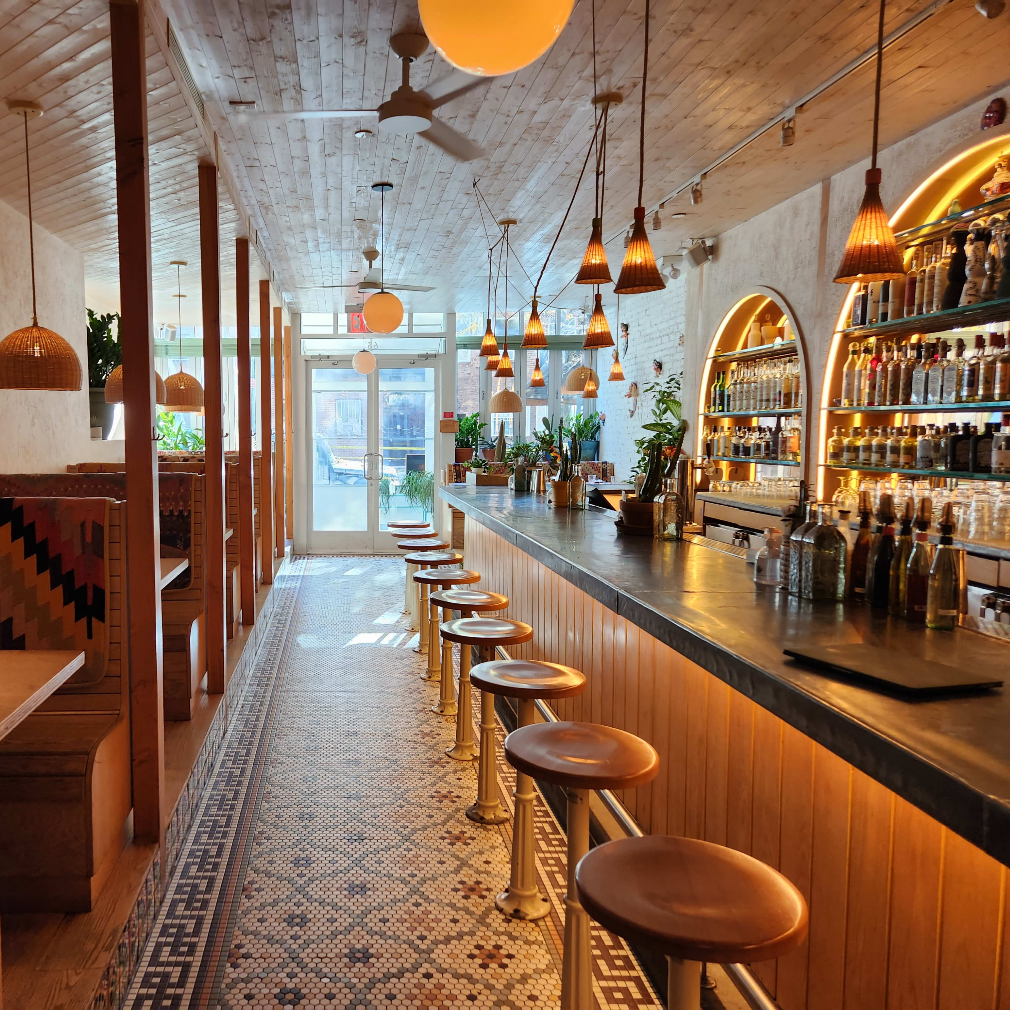 A modern restaurant interior with a long bar, wooden beams, and decorative lighting, featuring seating along one side and a well-stocked bar on the opposite wall.