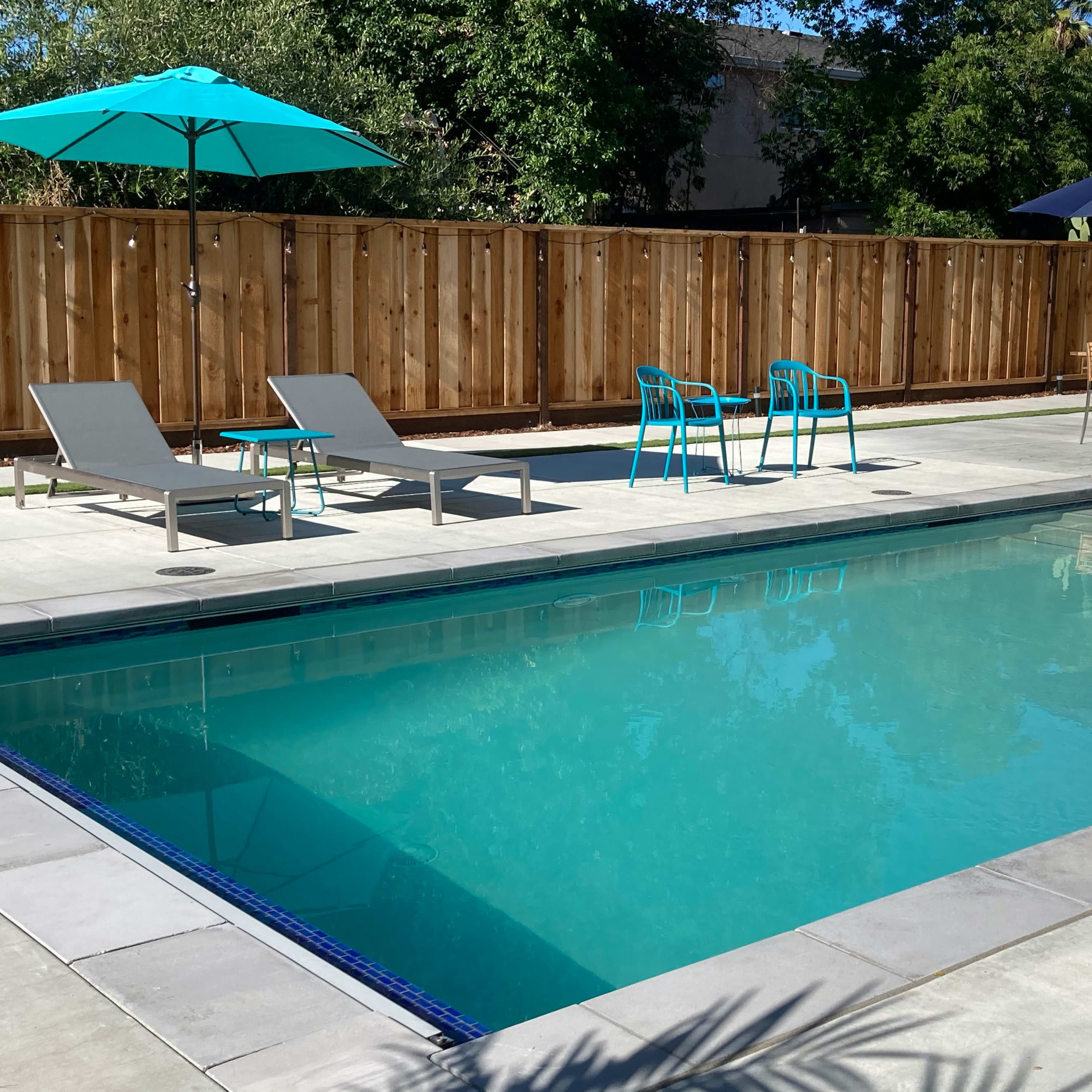 A clear swimming pool surrounded by lounge chairs, outdoor tables, and umbrellas in a fenced backyard.