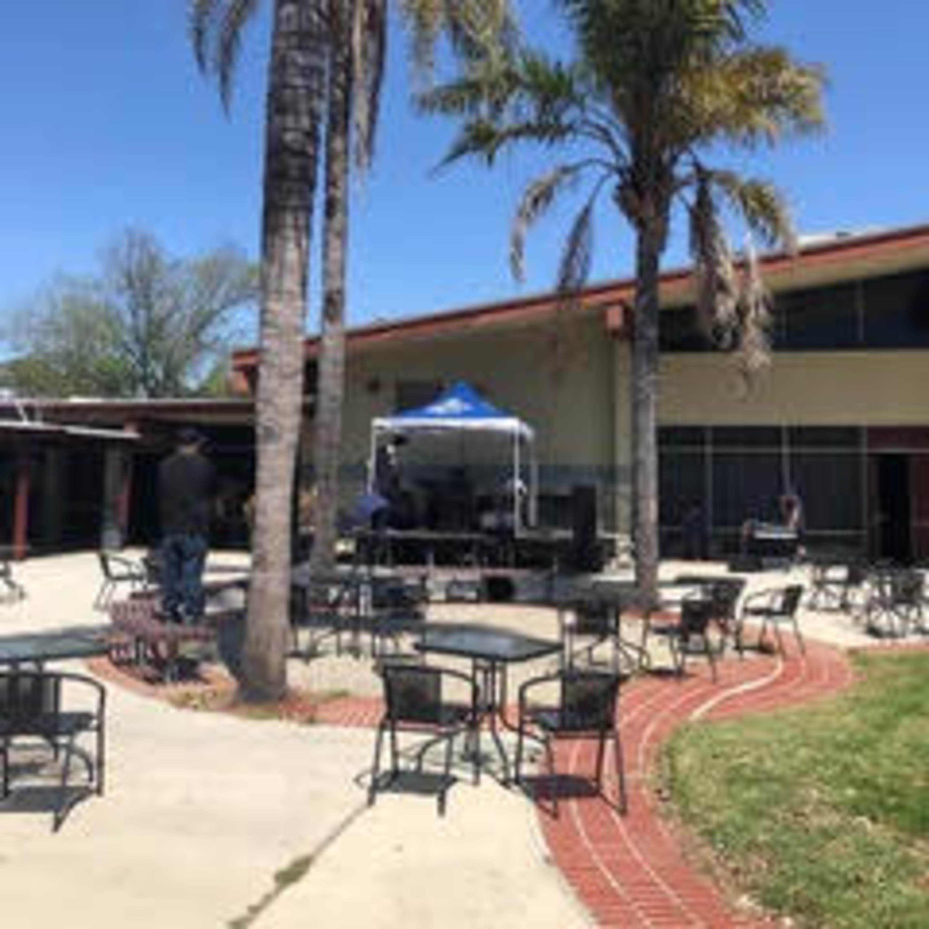 A stage with sound equipment is set up under a blue tent in a courtyard surrounded by palm trees and outdoor tables.