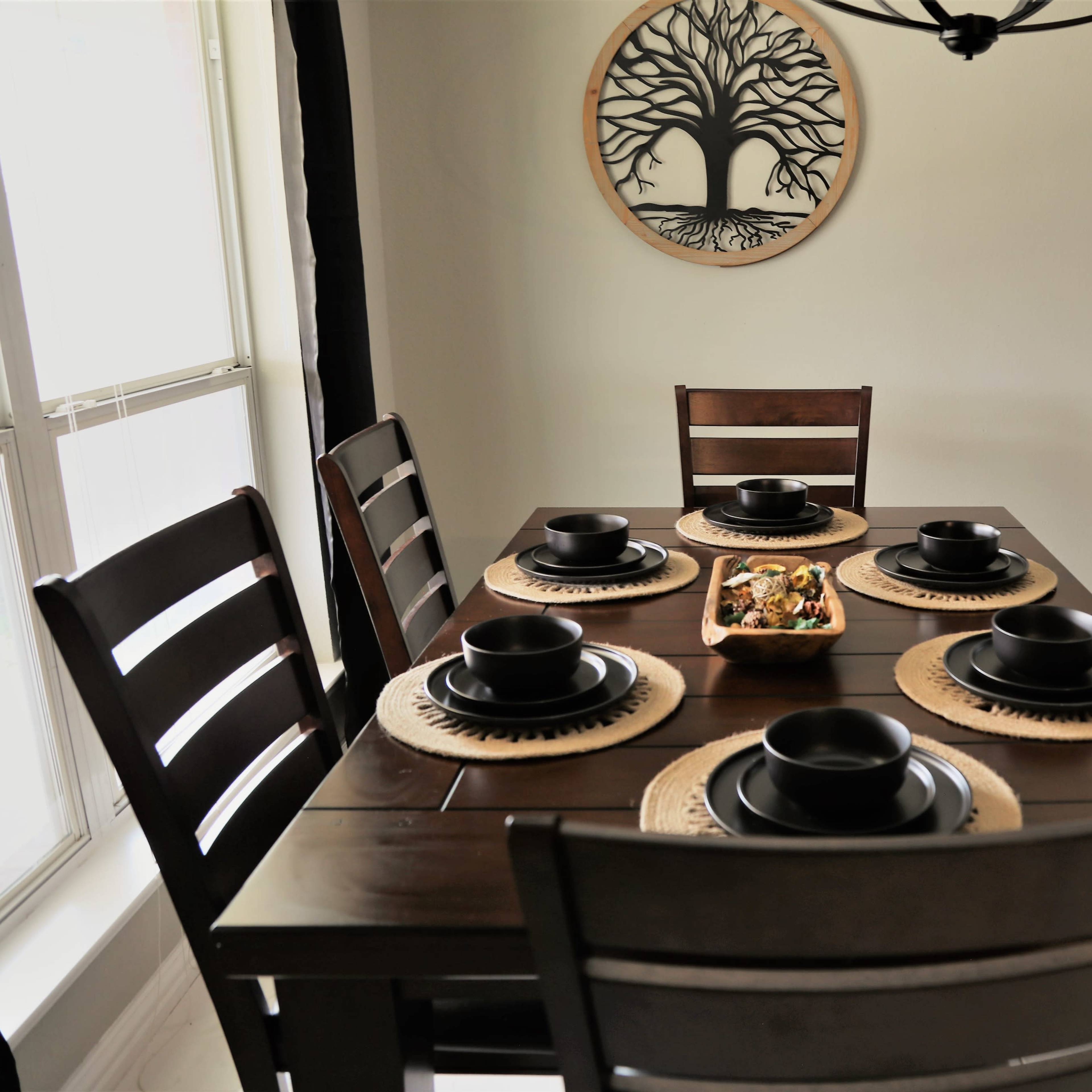 A wooden dining table is set with six black plates and bowls, surrounded by dark chairs, with a decorative tree wall art and snacks on a tray in the center.