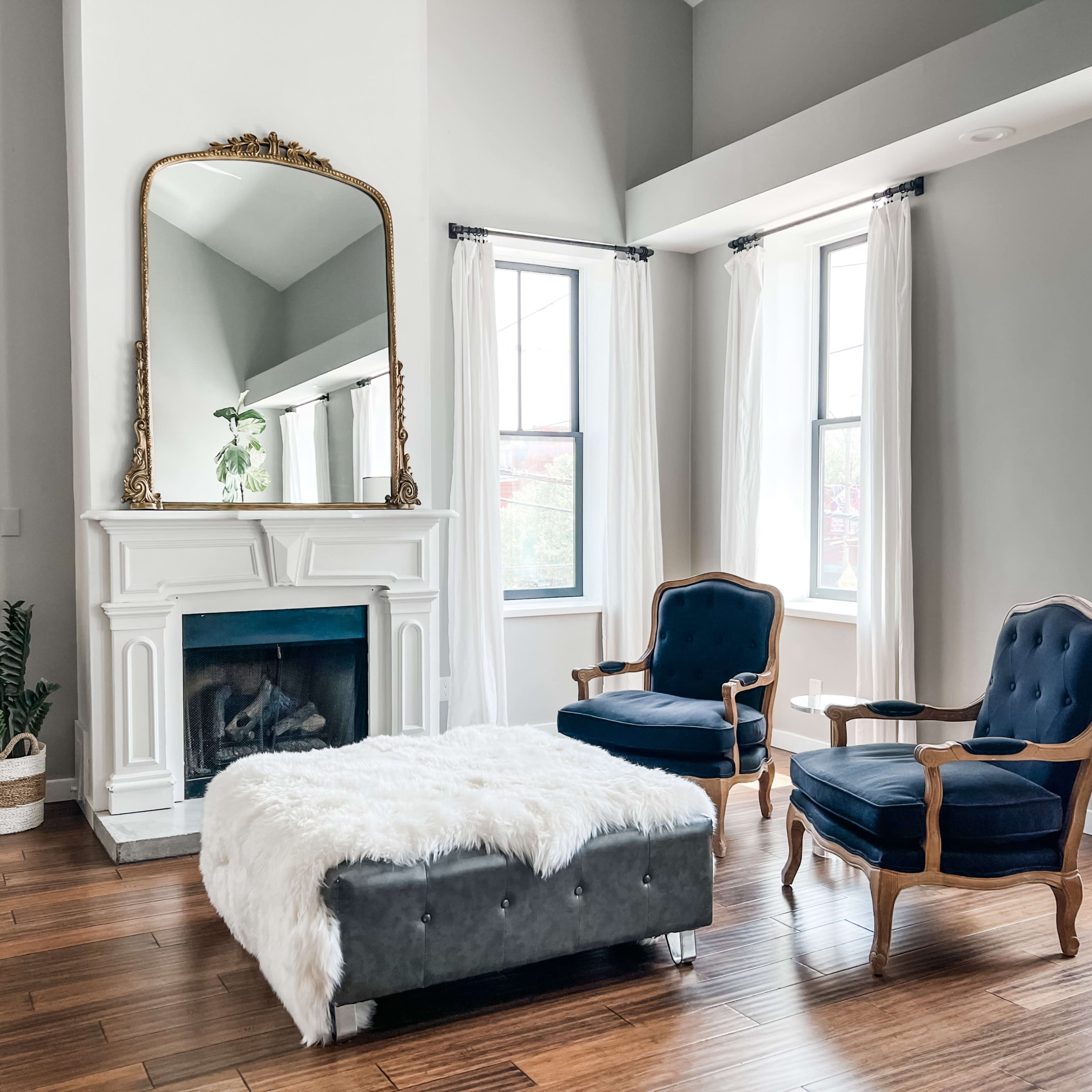 A living room with a white fireplace, a large mirror, two blue armchairs, and a fur-covered ottoman, all set against light gray walls and large windows.