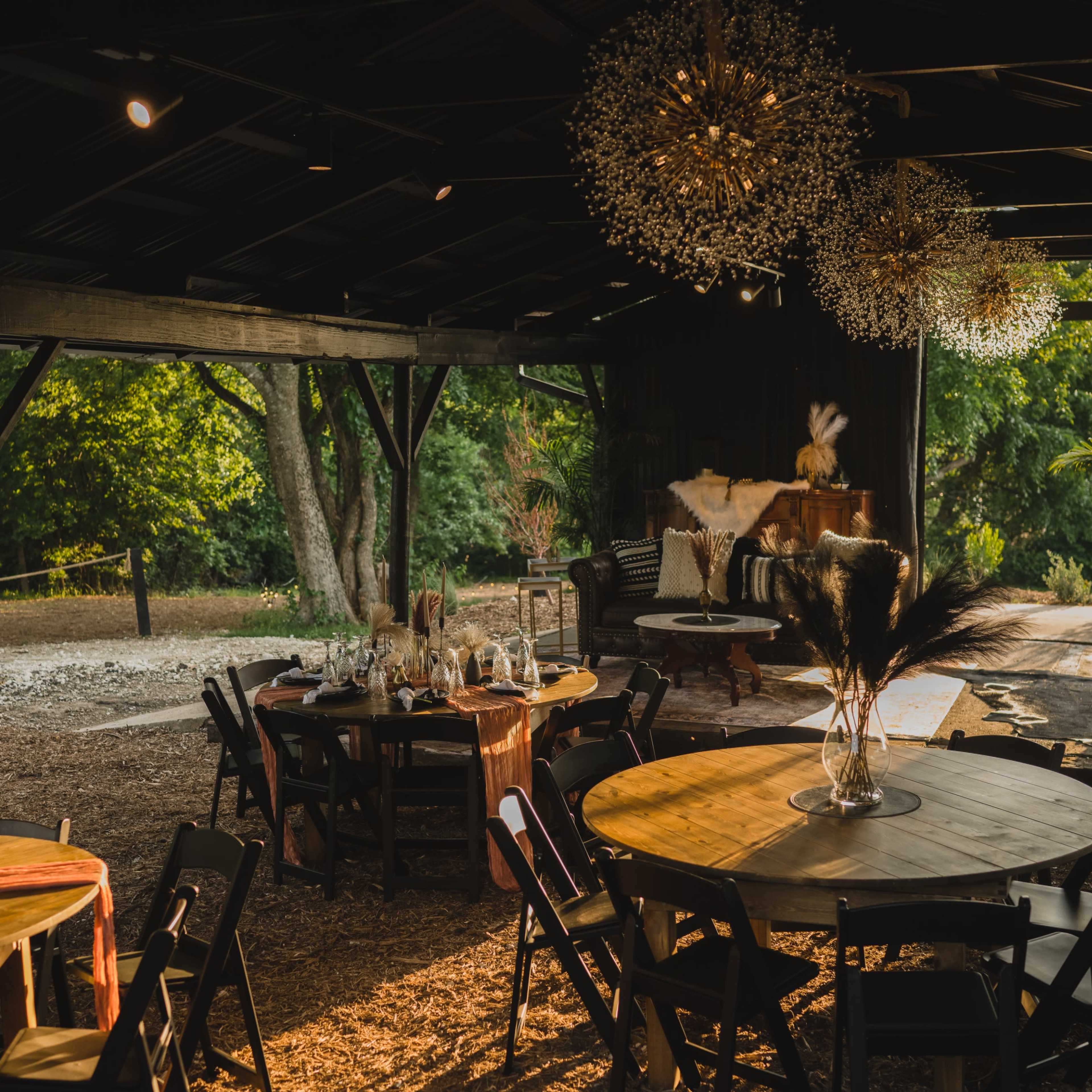 A rustic indoor venue with round tables set for an event, illuminated by decorative chandeliers and surrounded by greenery.