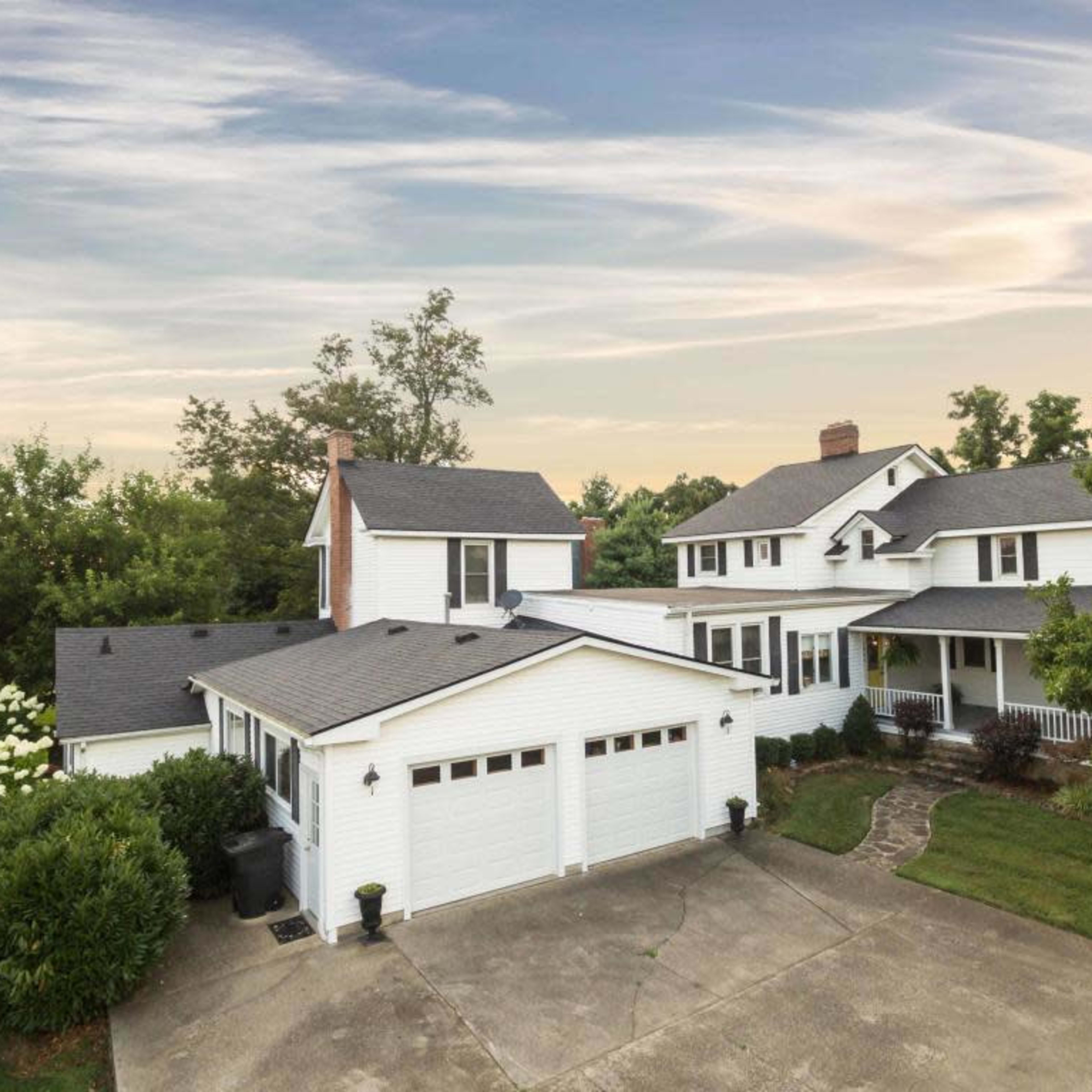 A white two-story house with multiple sections and a garage is surrounded by greenery and features a paved driveway.