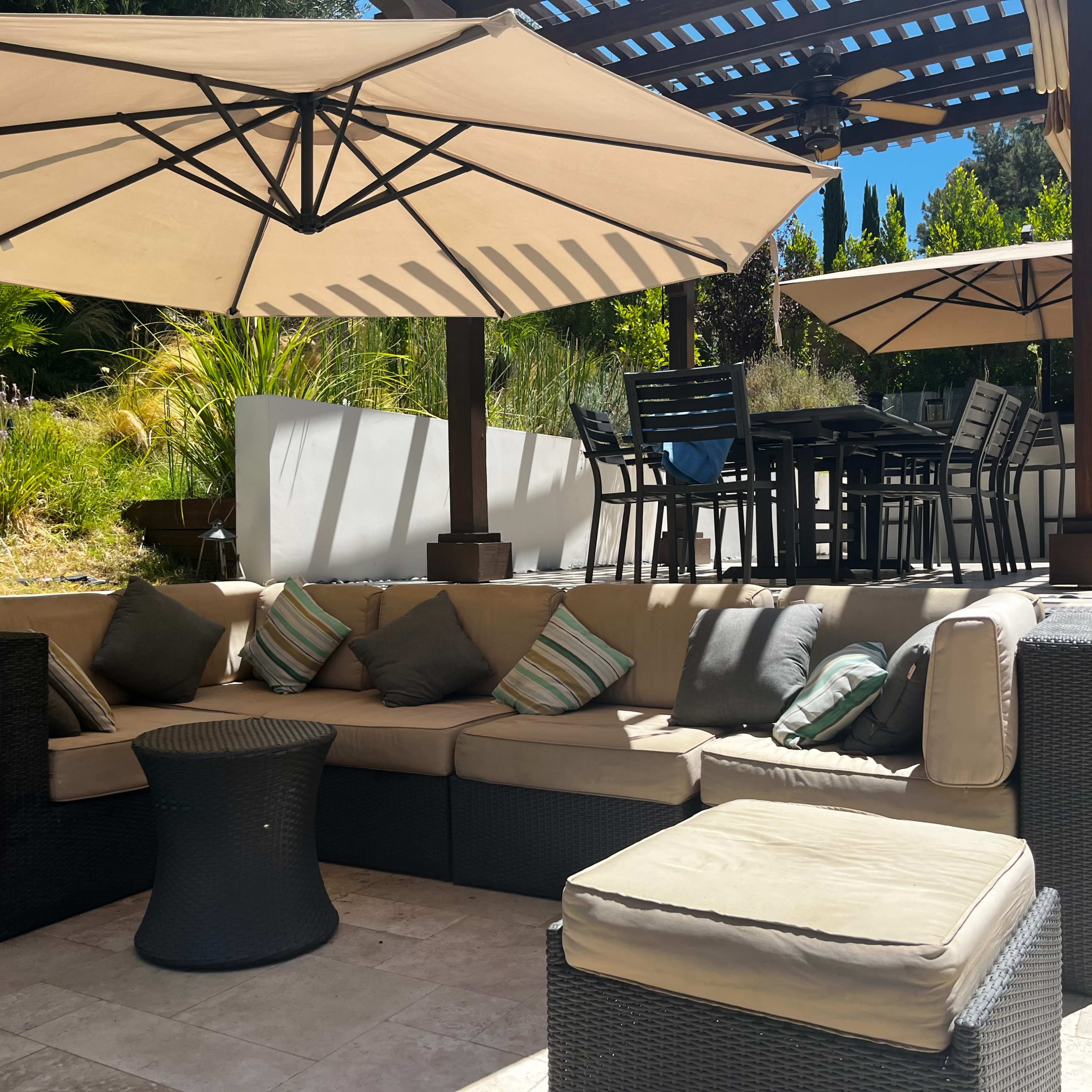 A shaded outdoor seating area features a sectional sofa with decorative cushions, a small round table, and a dining set under large umbrellas.