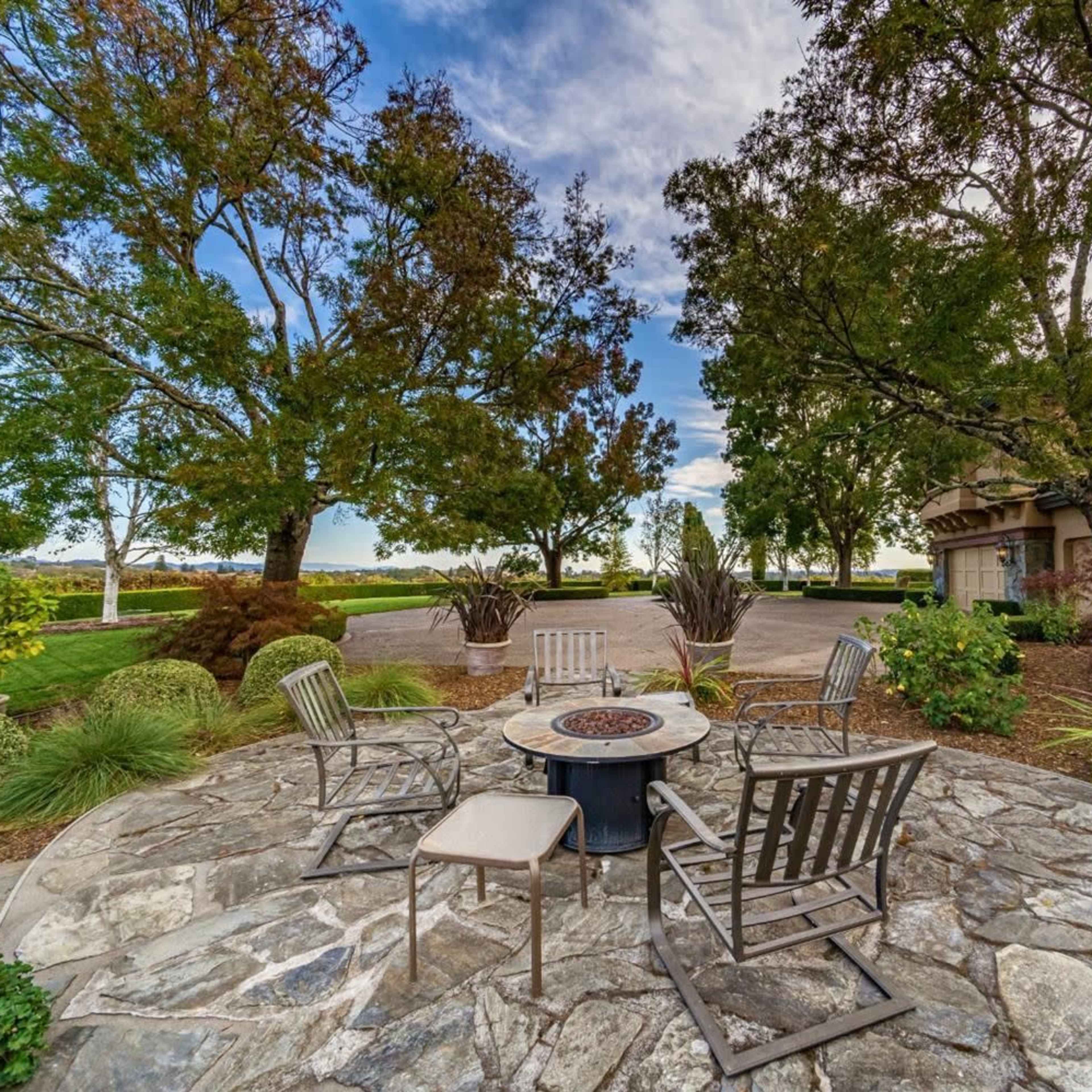 A stone patio features a circular fire pit surrounded by several metal chairs and lush greenery, with trees and a lake visible in the background.