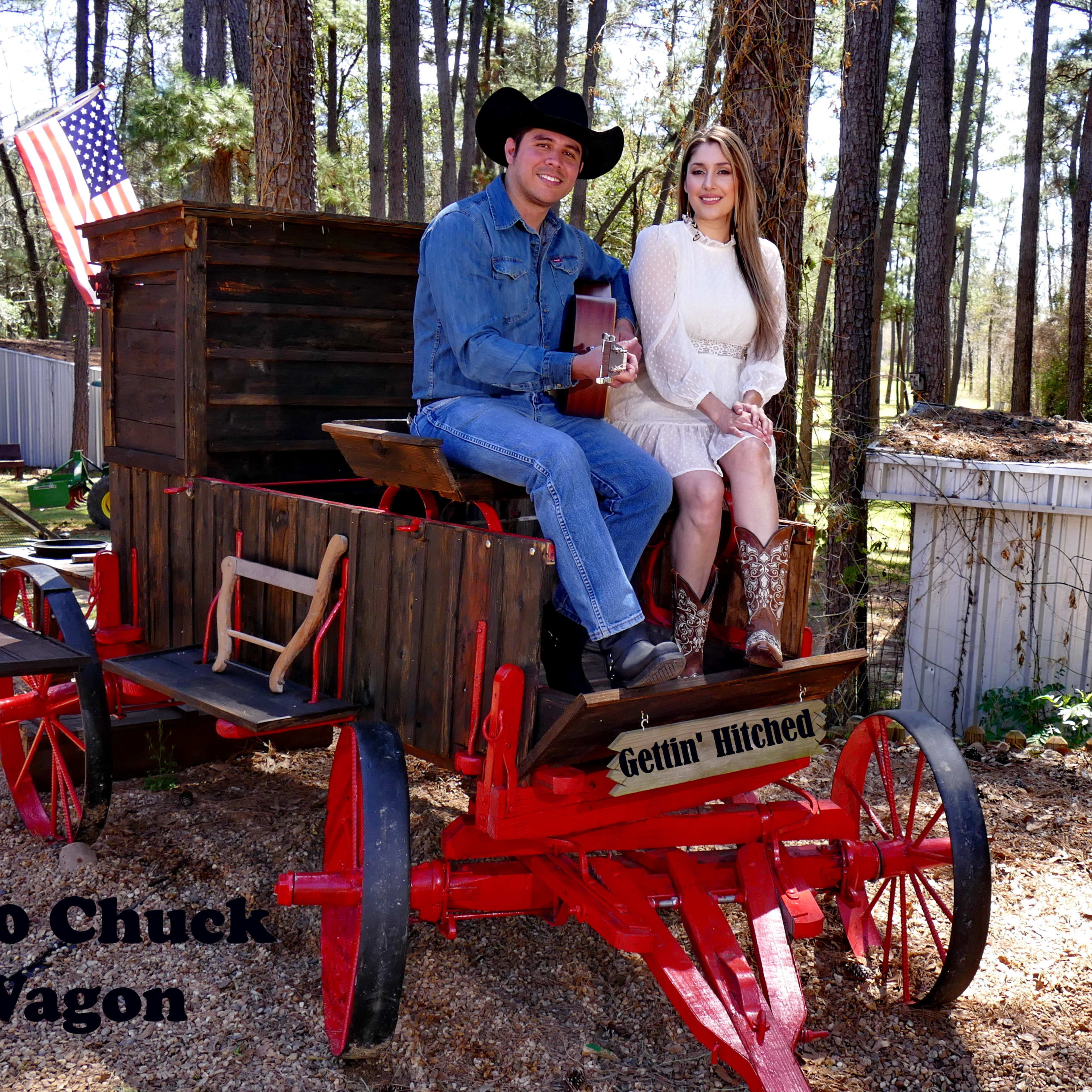 A man and a woman sit together on an antique red chuck wagon in a wooded area, with an American flag in the background.