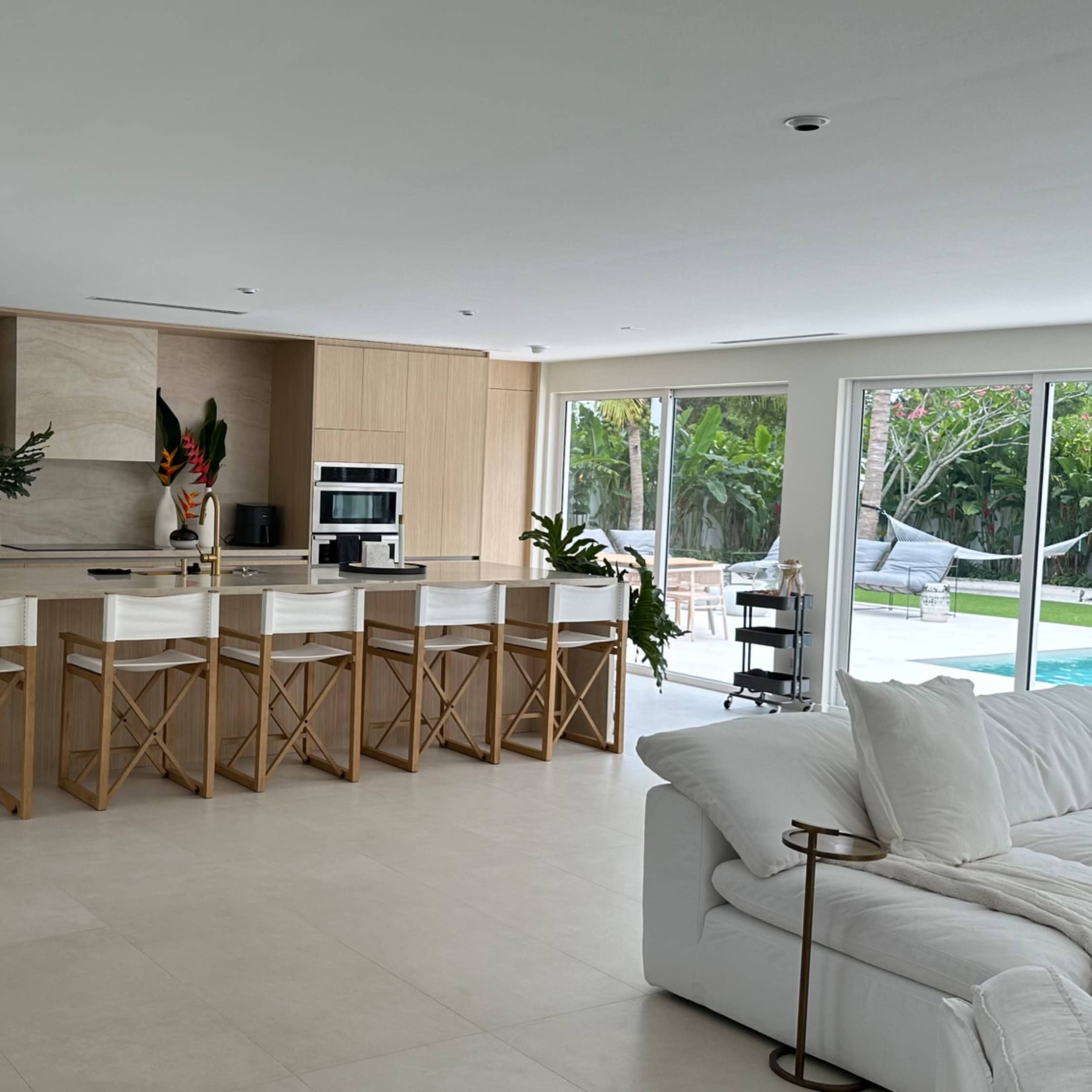 A modern kitchen with an island and bar seating, surrounded by large windows overlooking a pool area.