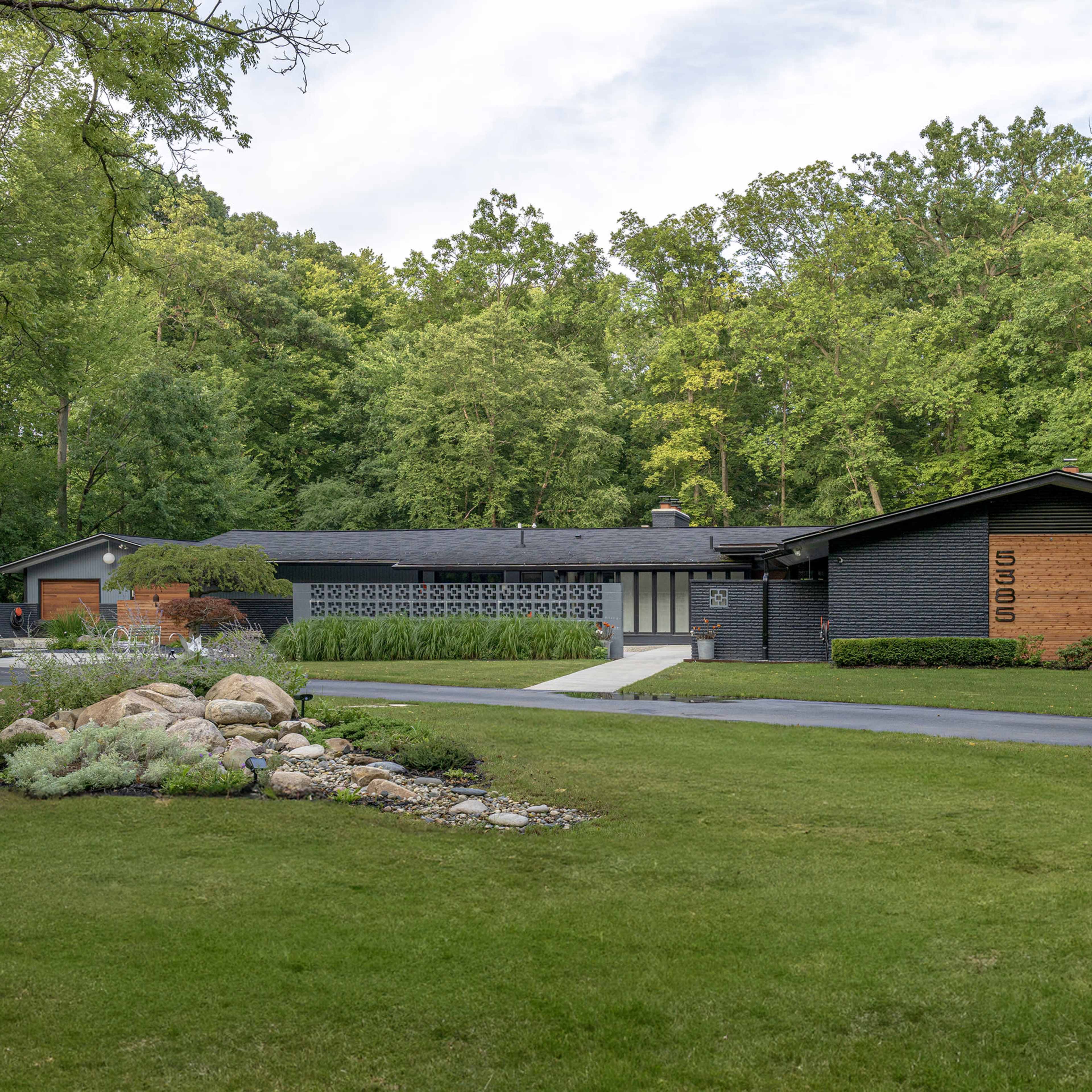 A modern black house set in a wooded area, with a neatly landscaped yard and a curved driveway leading up to the entrance.