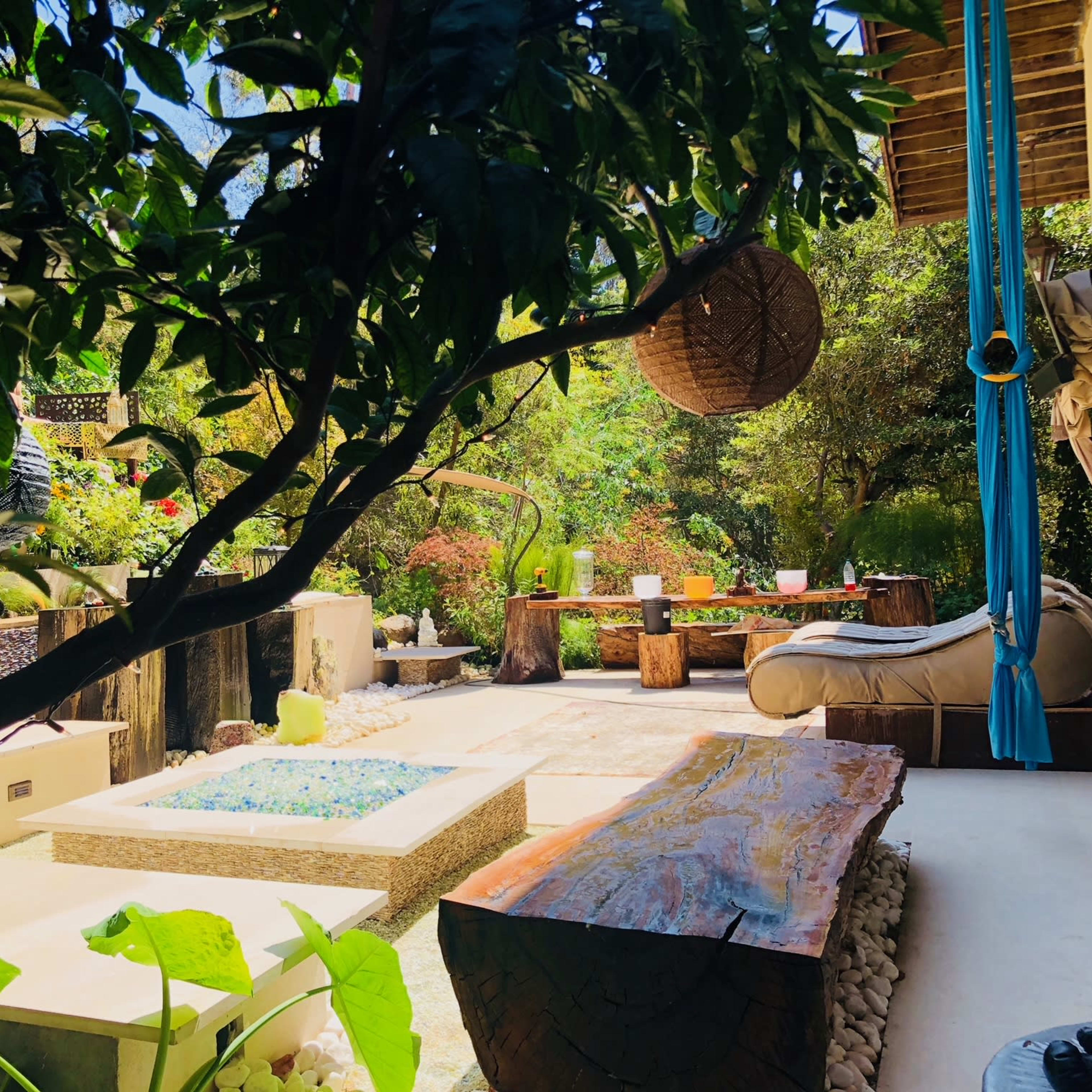 A lush outdoor garden area featuring a small pool, wooden seating, and a large wooden table surrounded by greenery.