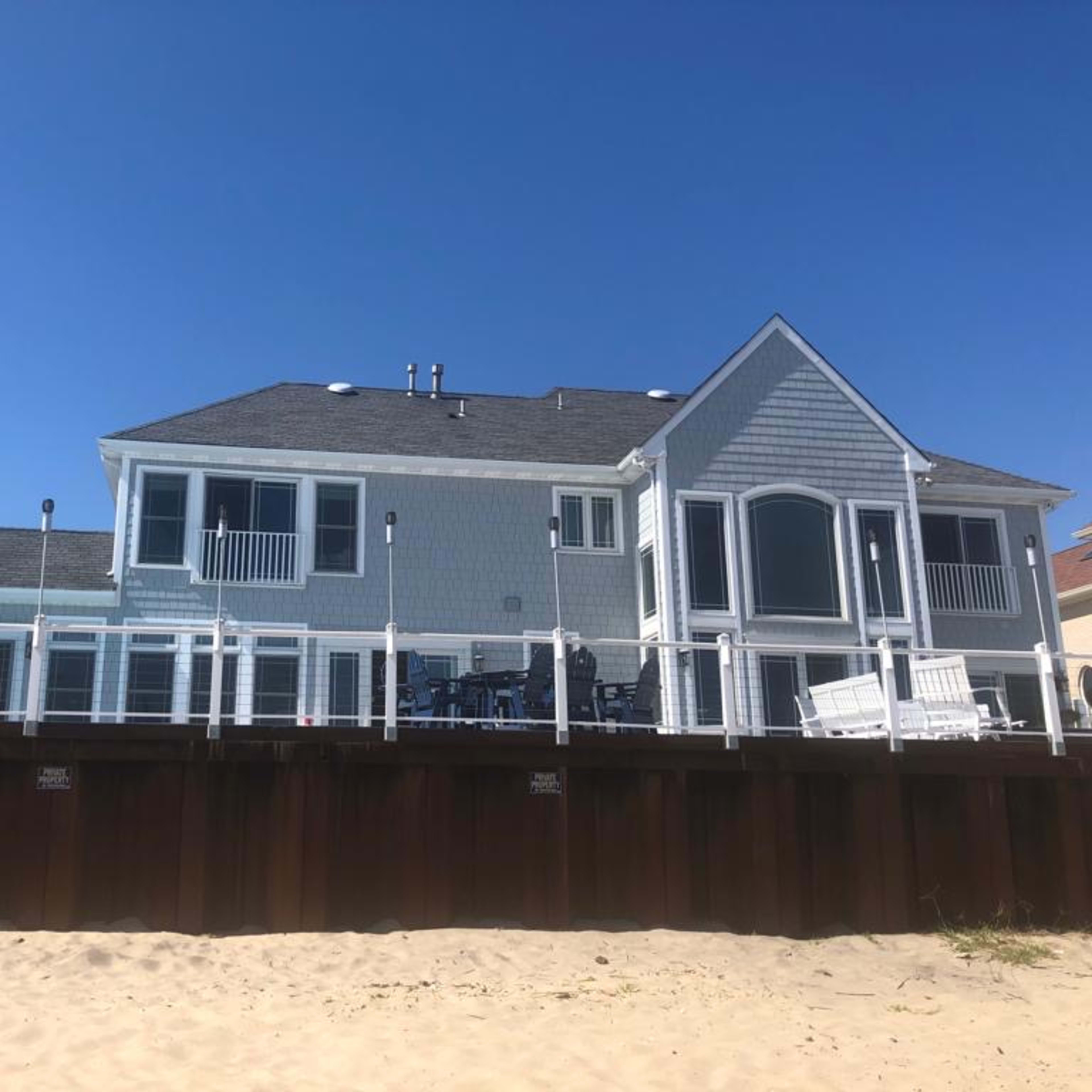 A beach house with a gray exterior and a wooden deck overlooks a sandy shore under a clear blue sky.