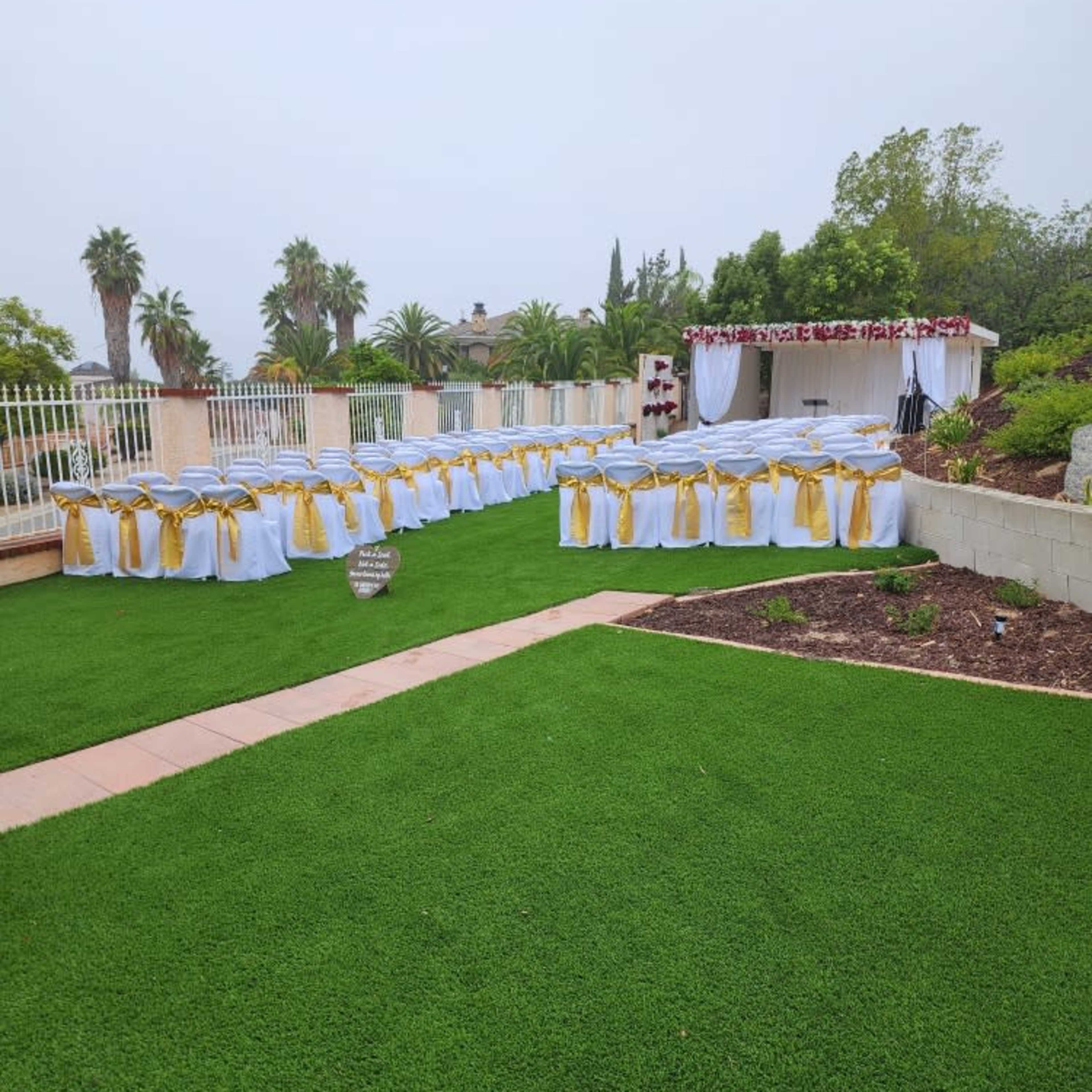 A beautifully arranged outdoor event space features tables dressed in white tablecloths with gold sashes, set up on vibrant green grass.
