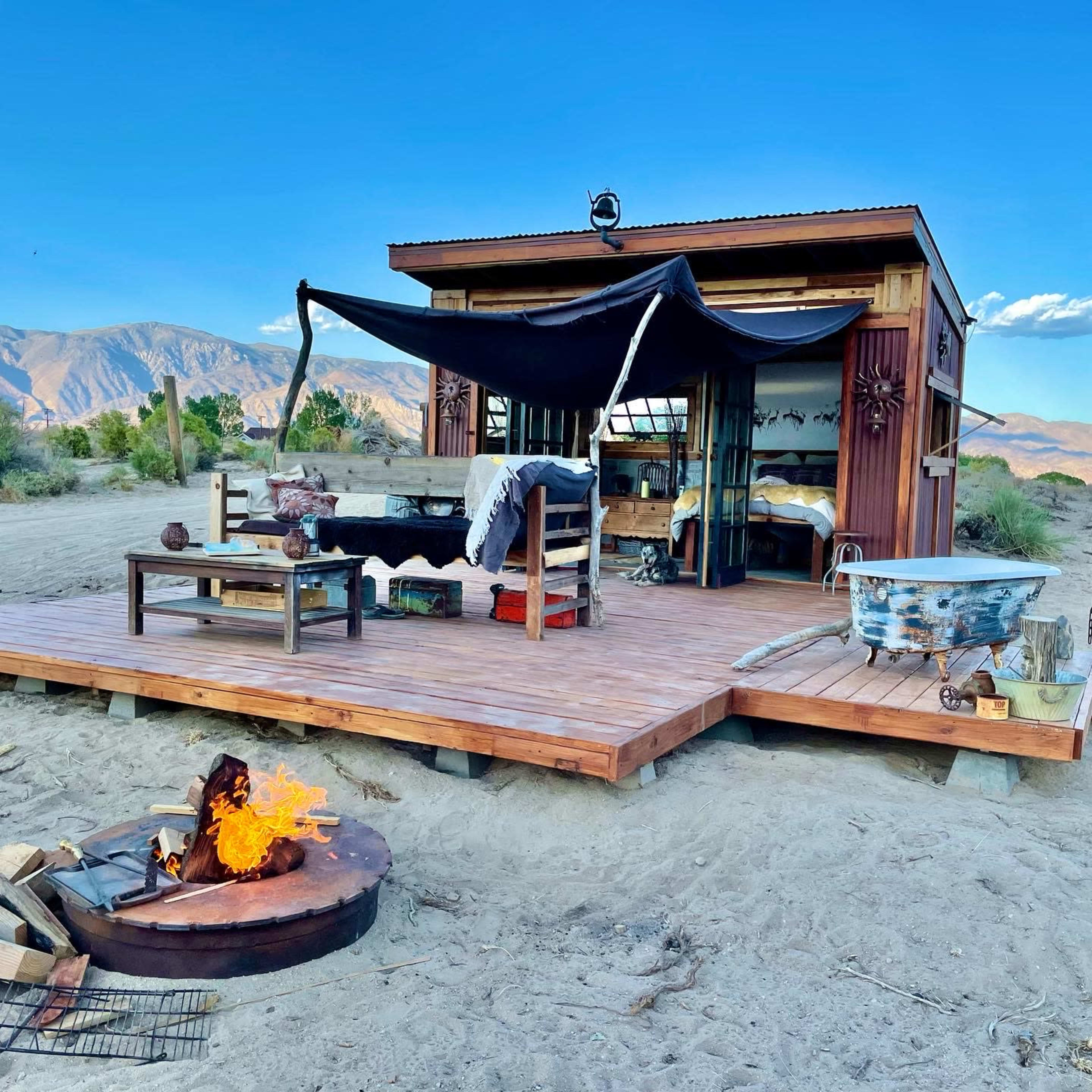 A small wooden cabin with a shaded porch and a fire pit sits in a desert landscape surrounded by mountains.