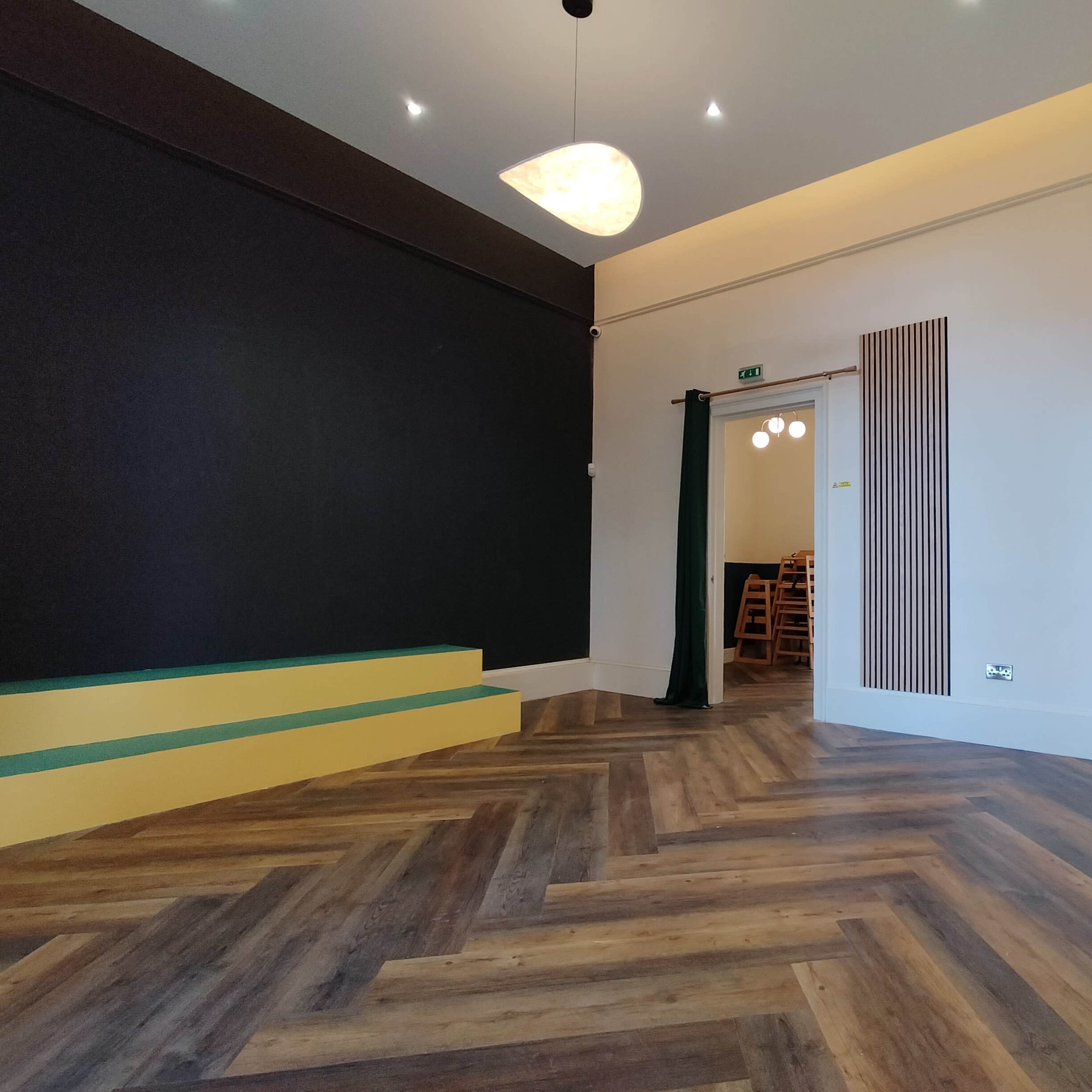 A spacious interior room with dark walls, a wooden chevron floor, a ceiling light fixture, and a doorway leading to a dining area with wooden chairs visible.