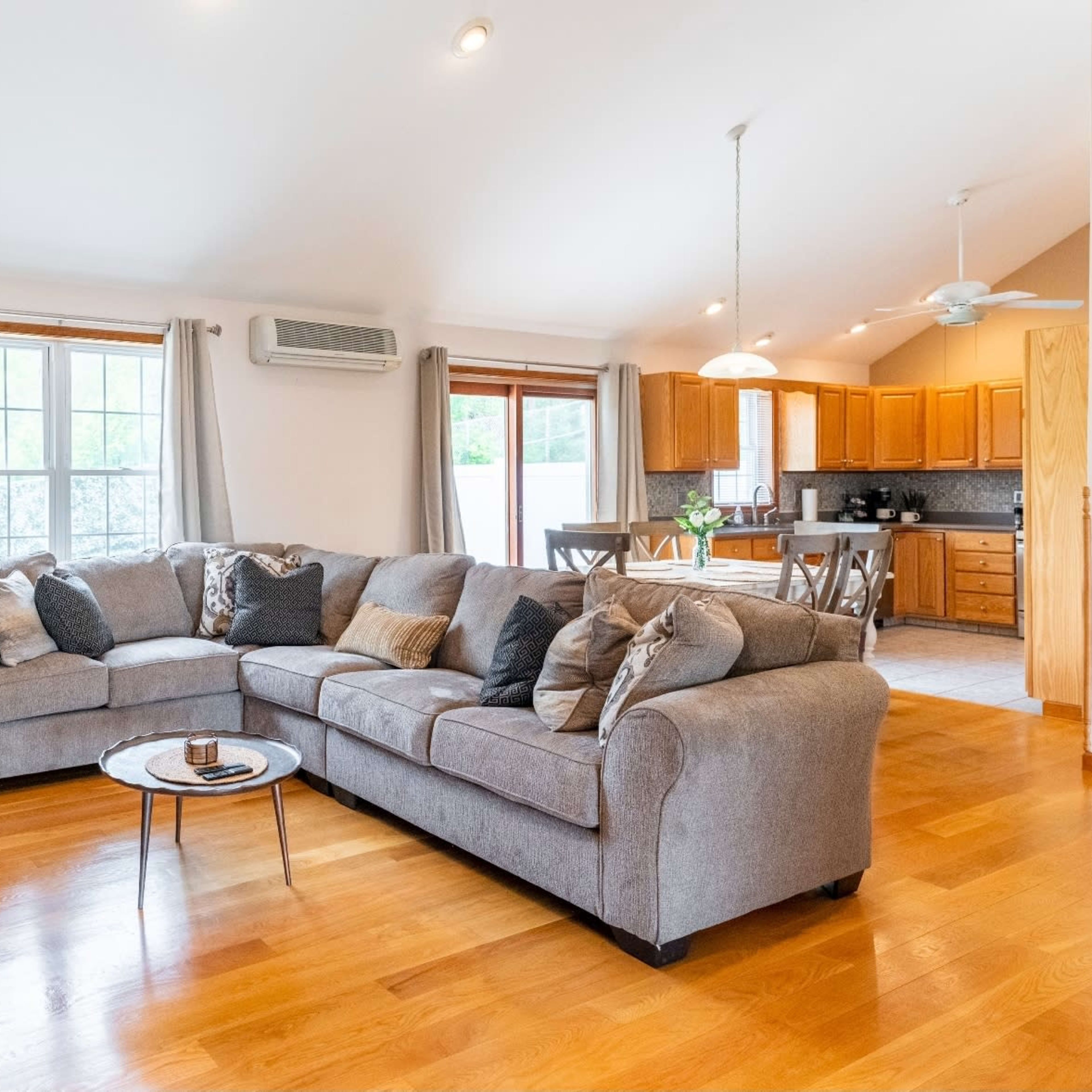 A spacious living room features a large gray sectional sofa, wooden floors, and an open layout leading to a kitchen with wooden cabinetry.