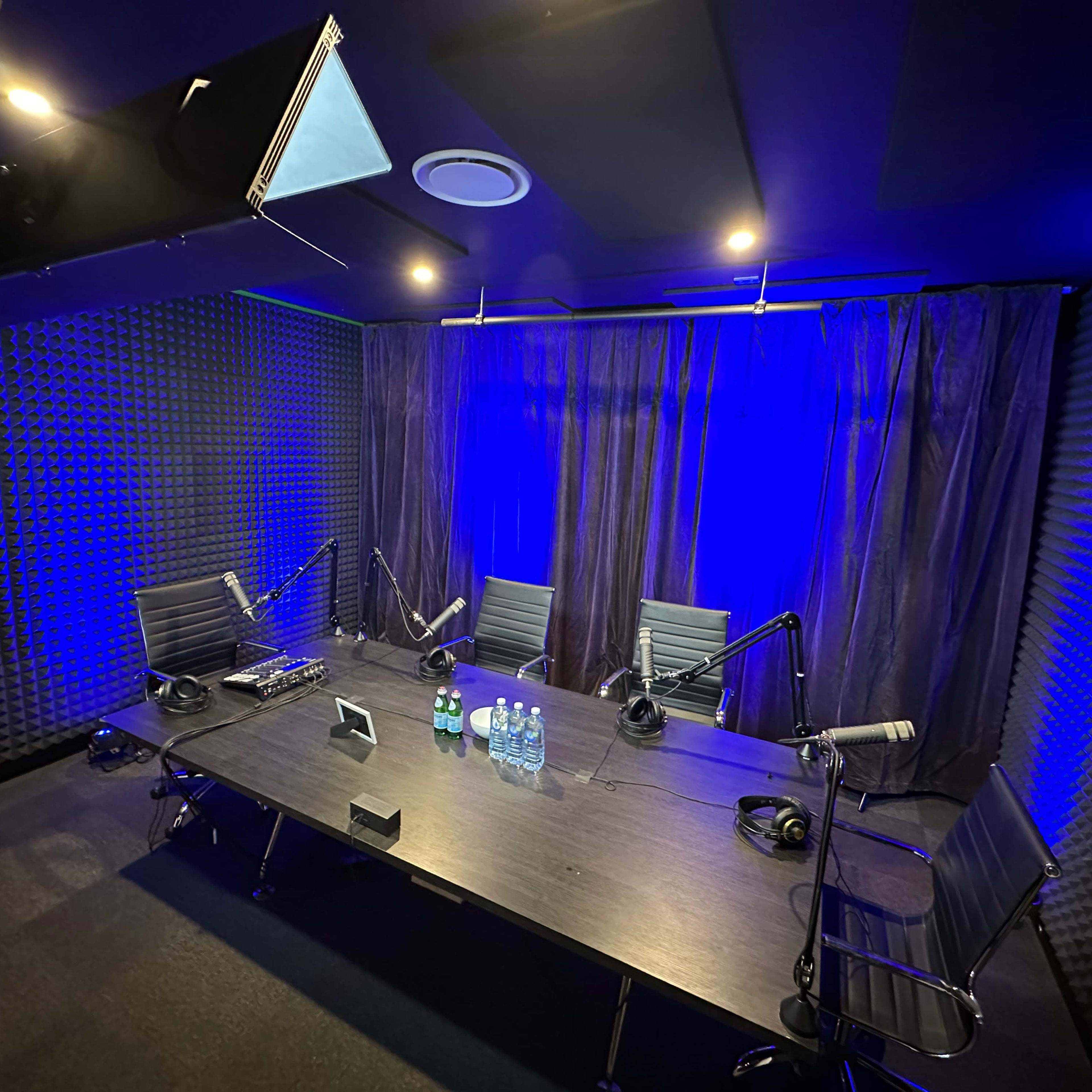A recording studio with a large table, four chairs, and multiple microphones, surrounded by soundproof walls and blue lighting.
