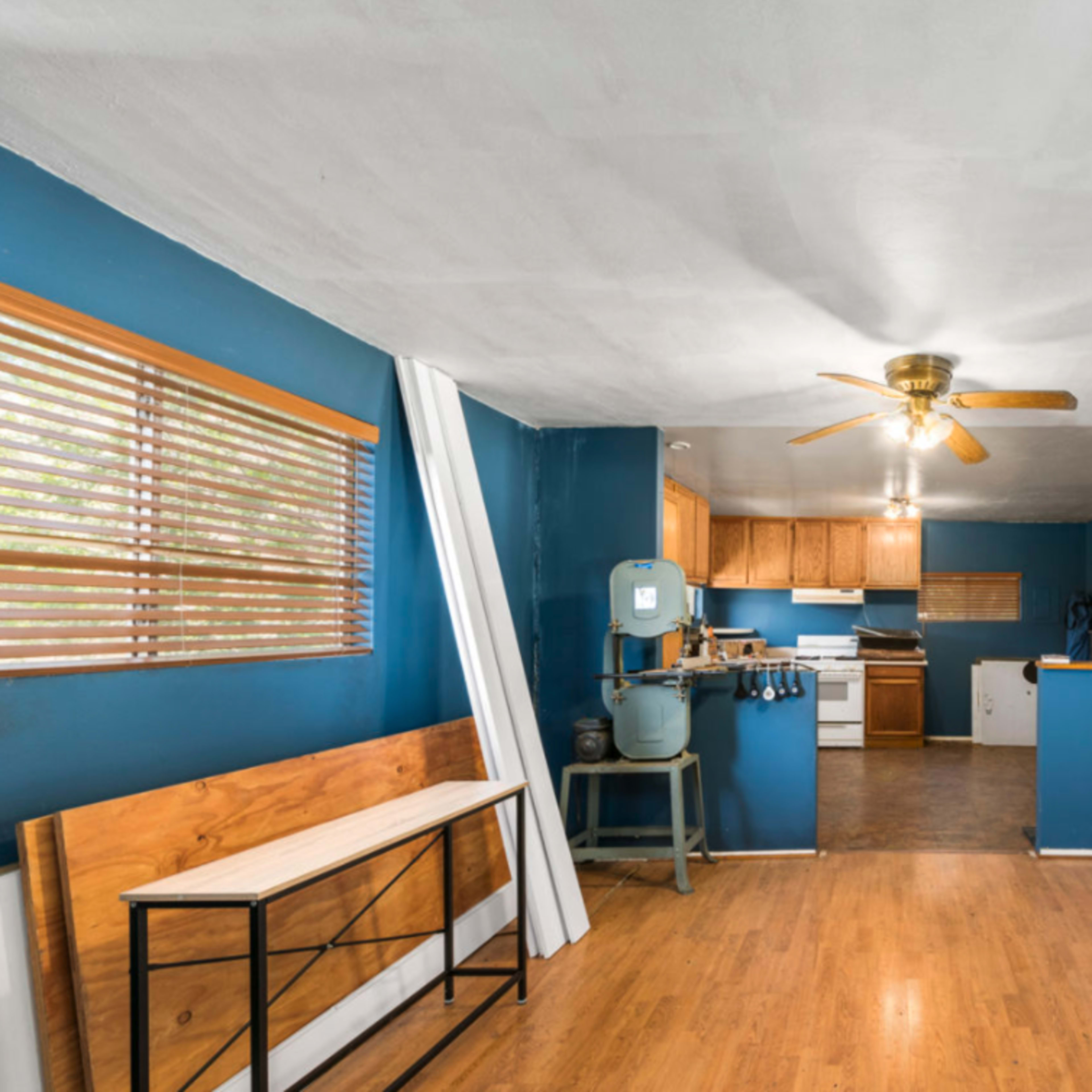 A room with blue walls, wooden flooring, and a ceiling fan, featuring a kitchen area with wooden cabinets and a white stove.