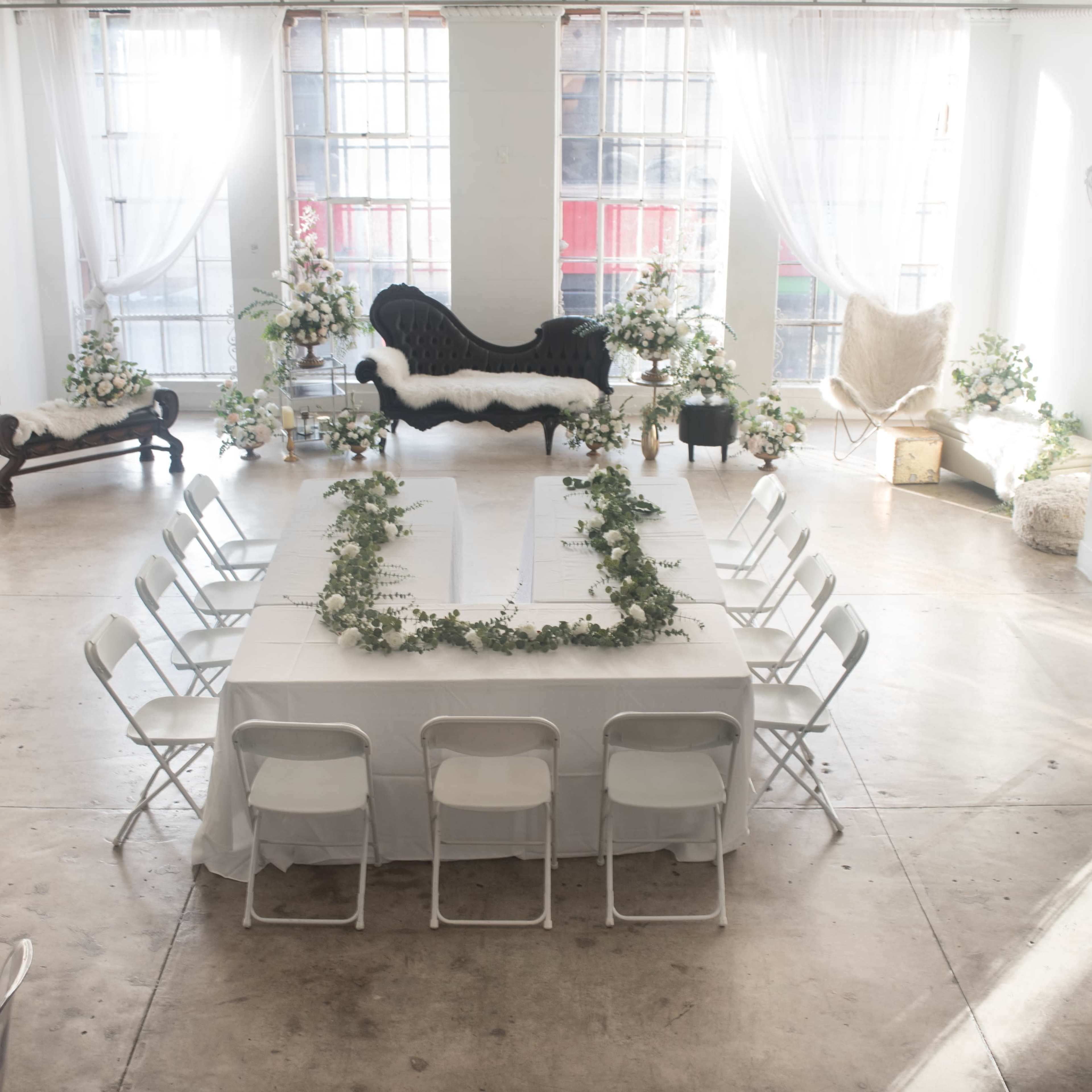 A bright event space arranged with a long table surrounded by chairs, decorated with floral arrangements and elegant furnishings.