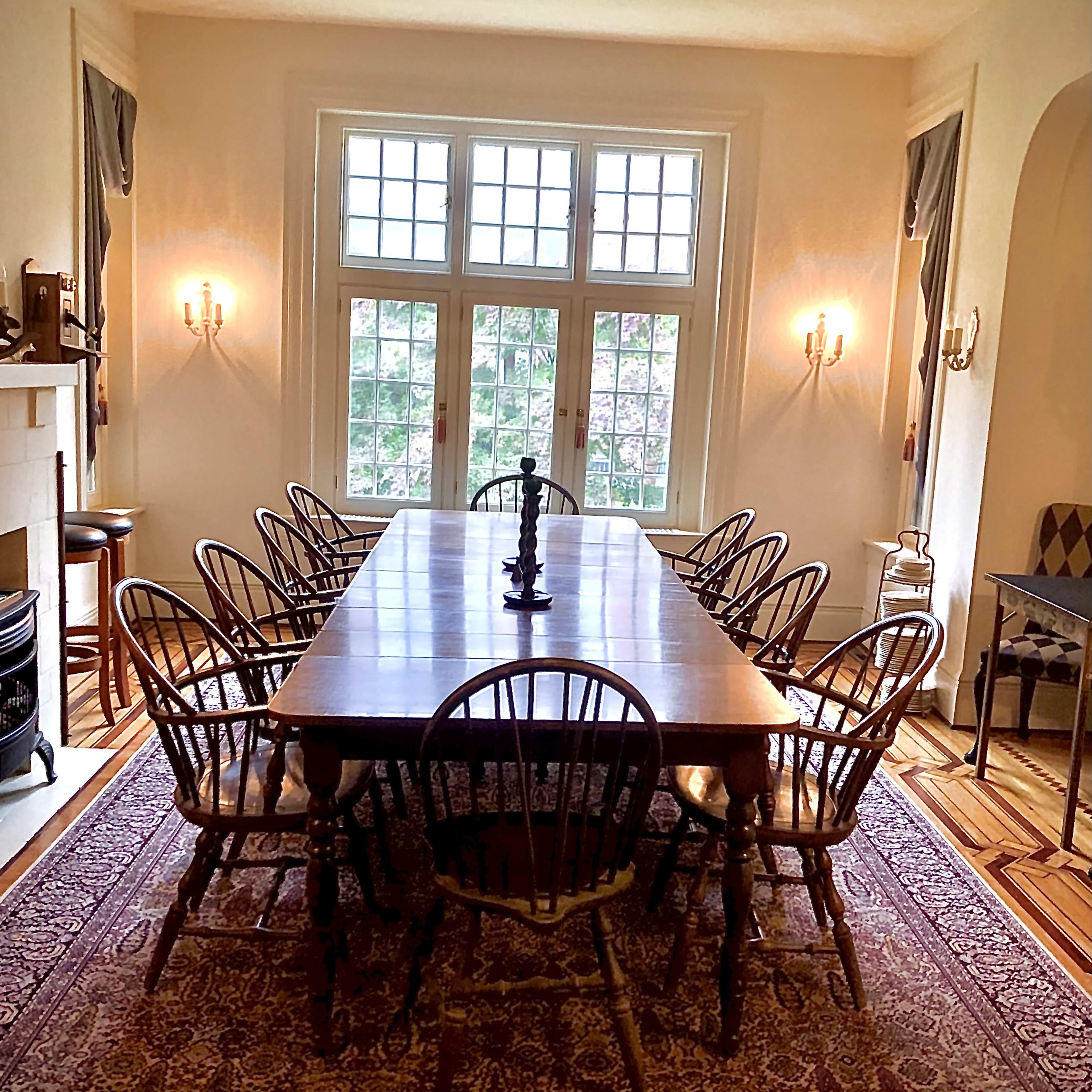 A large wooden dining table surrounded by ten chairs is positioned in a well-lit room with multiple windows and a decorative rug on the floor.