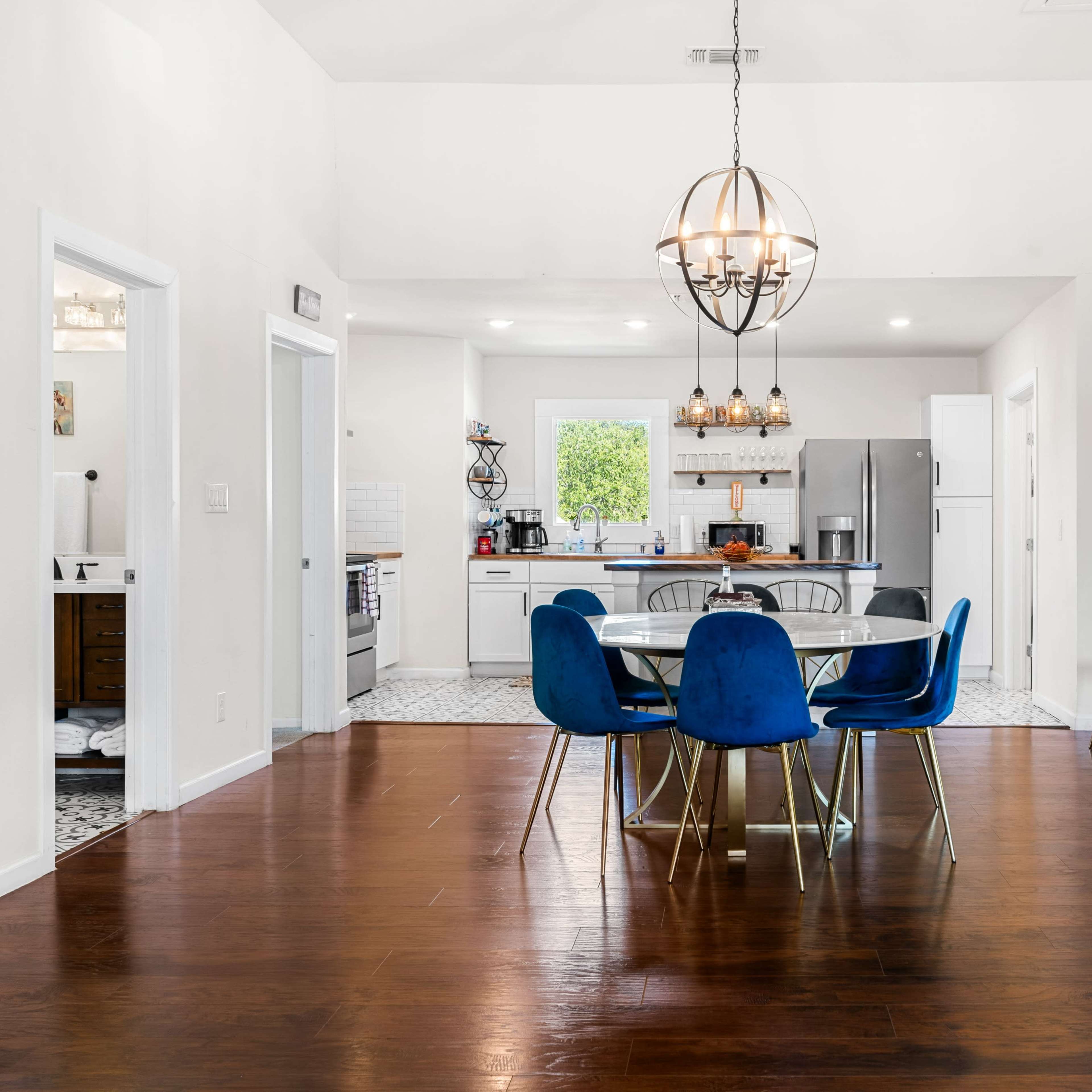 A bright dining area features a round table surrounded by blue chairs, with a view of a modern kitchen in the background.