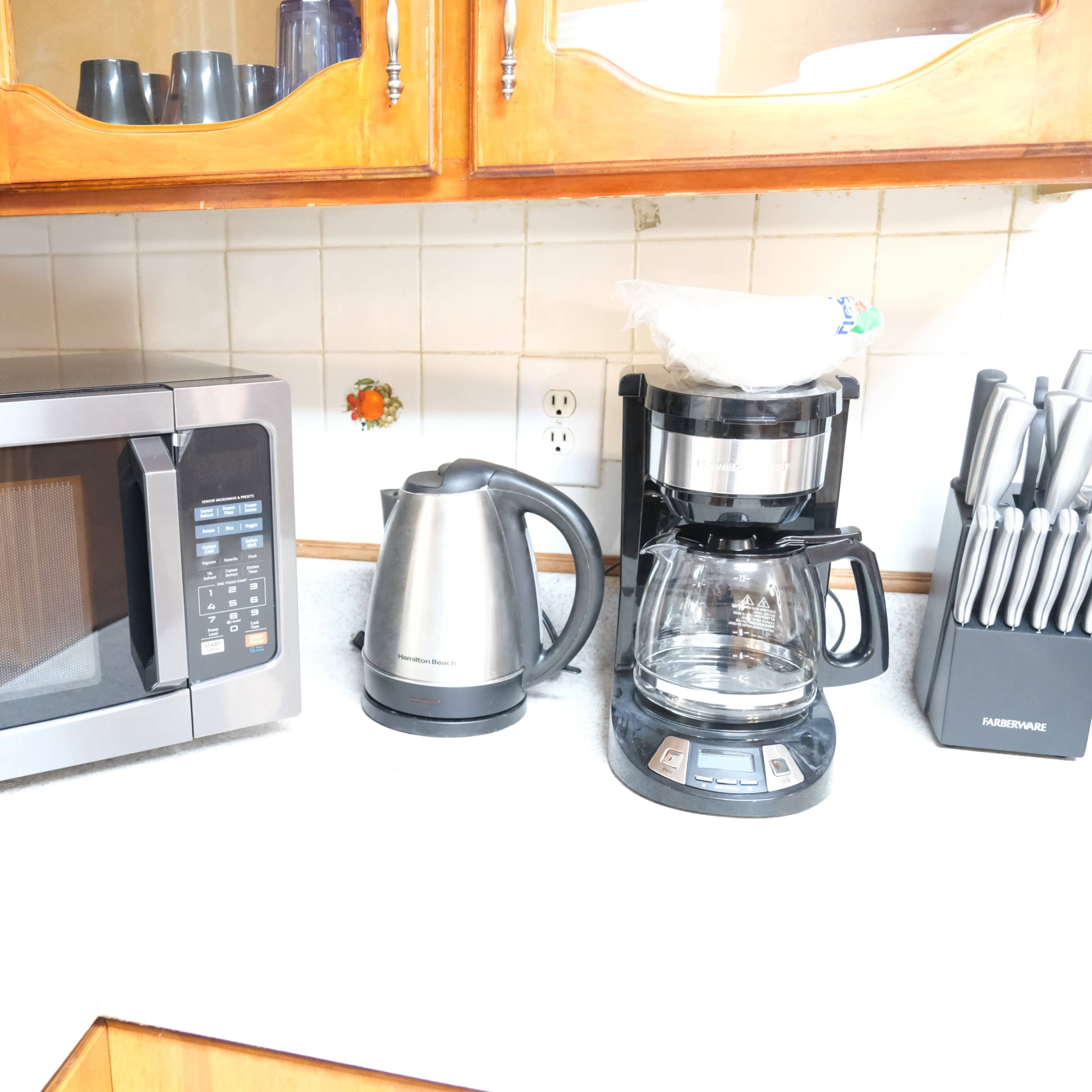 A kitchen countertop with a microwave, electric kettle, coffee maker, and a set of knives in a holder.
