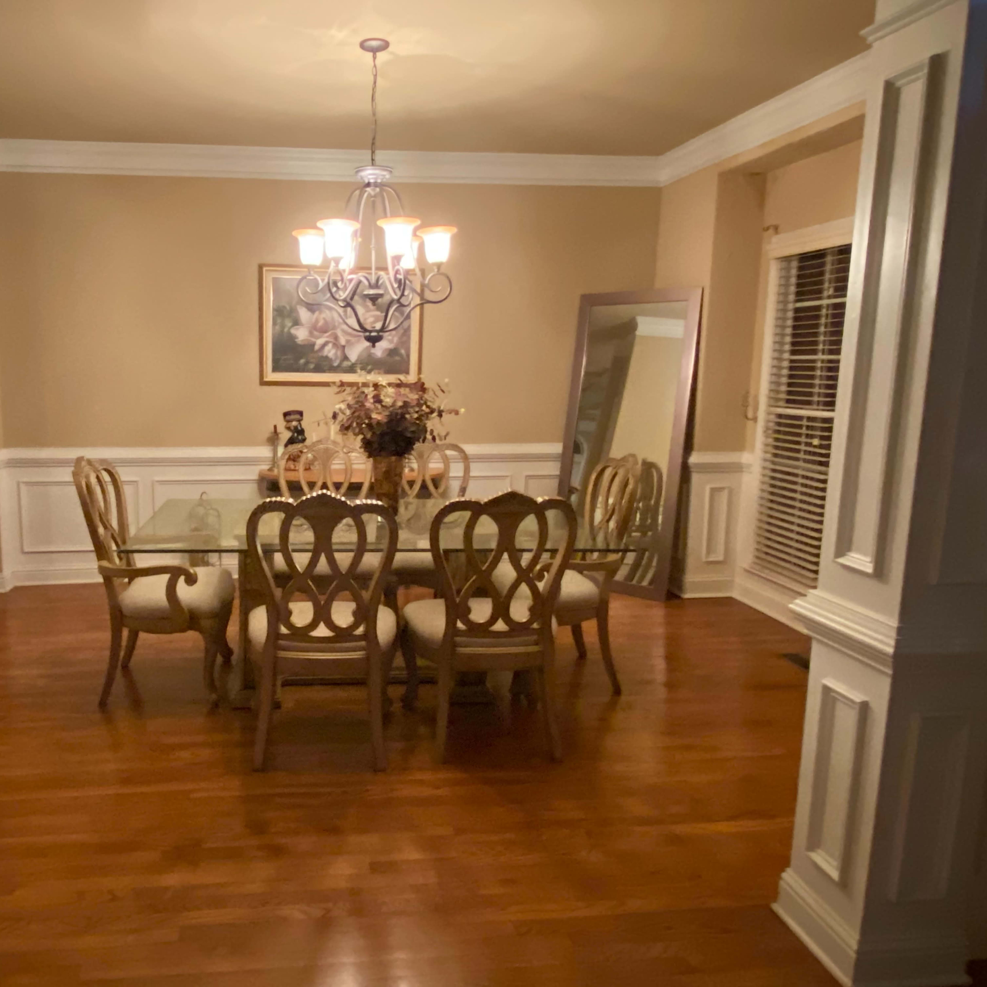 A dining room features a wooden table surrounded by chairs, illuminated by a chandelier, with a mirror and a framed painting on the walls.