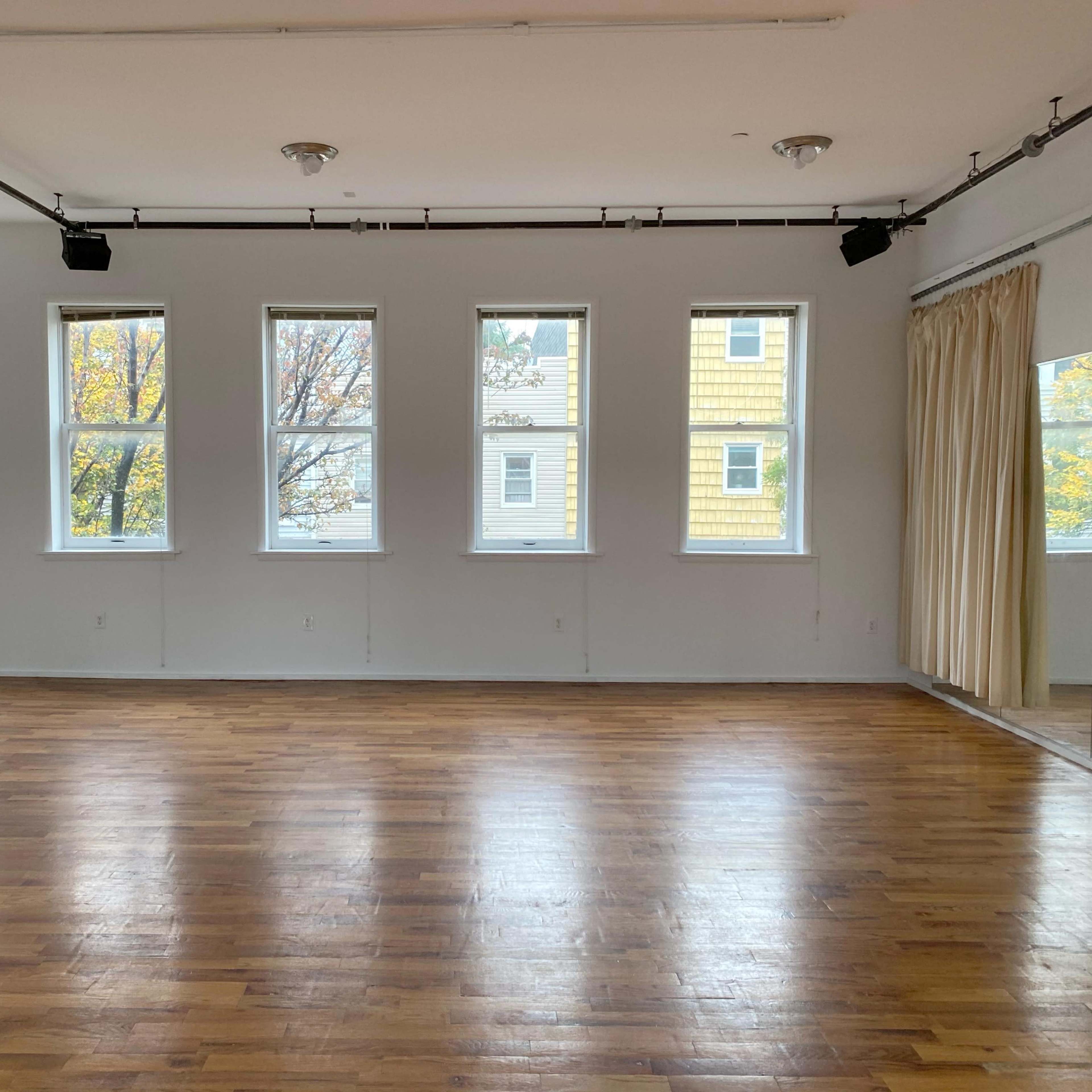 An empty wooden-floored room with large windows and a mirrored wall.