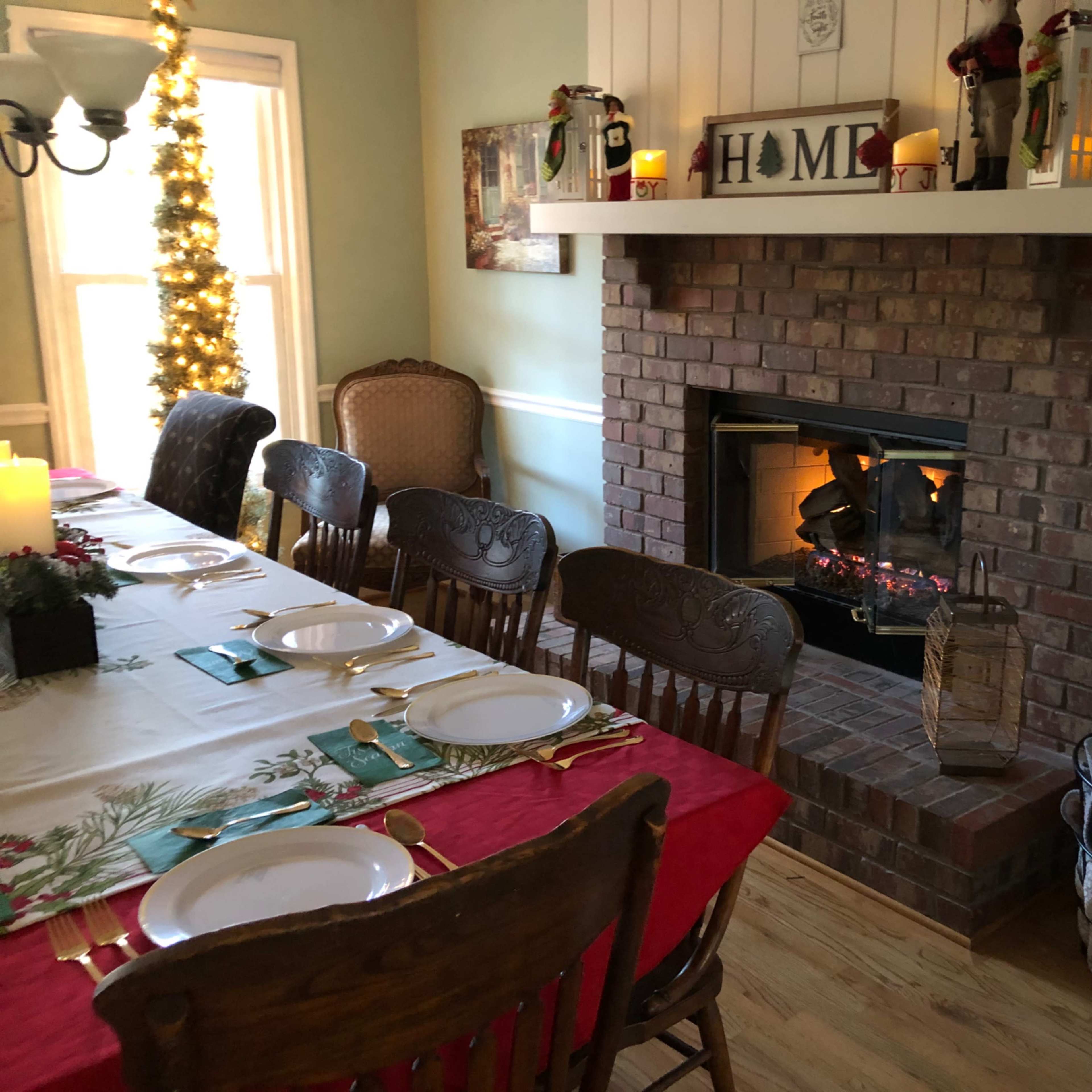 A dining area features a long table set for a meal, with a brick fireplace adorned with candles and holiday decorations.