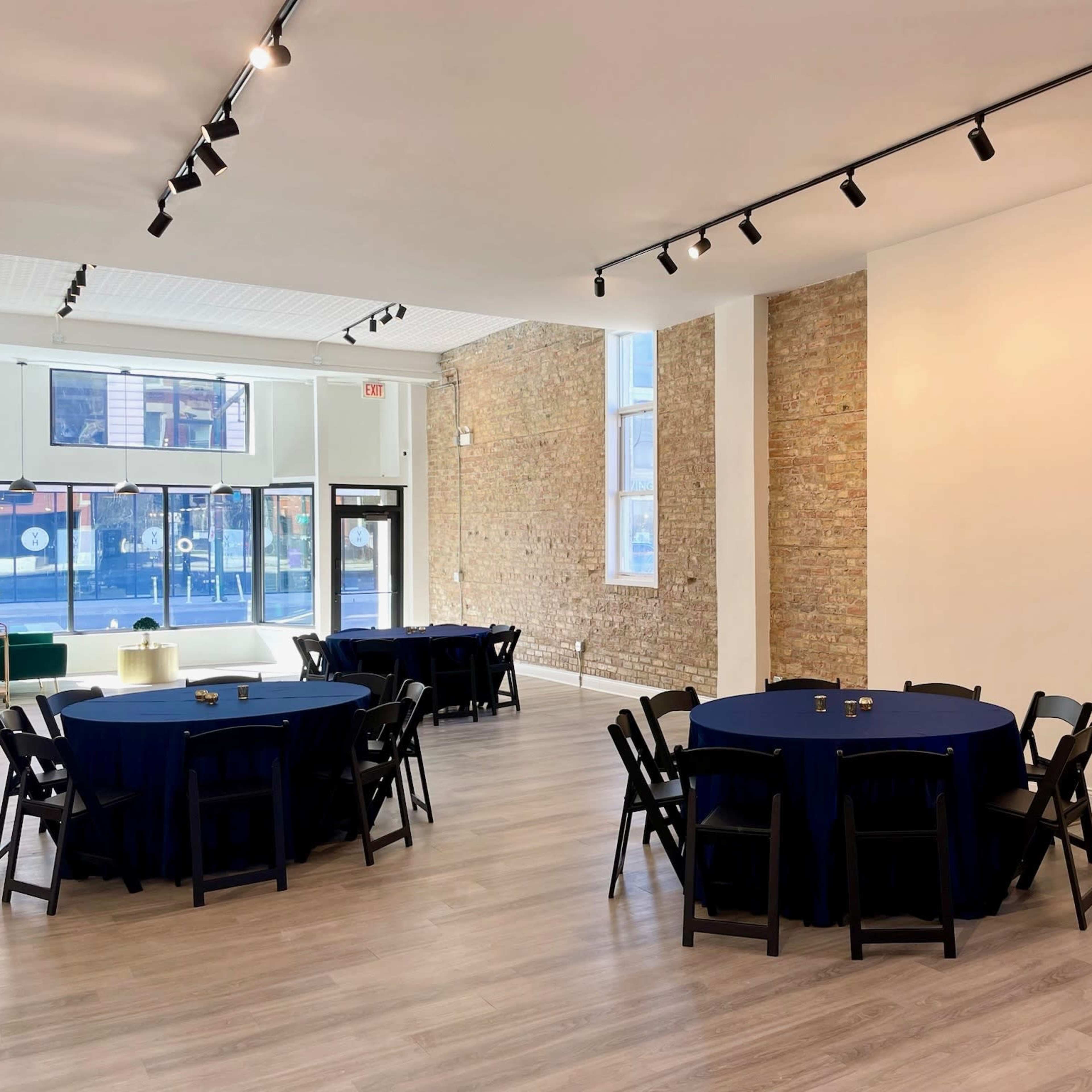 An interior space with two round tables covered in blue tablecloths, black chairs, and exposed brick walls.
