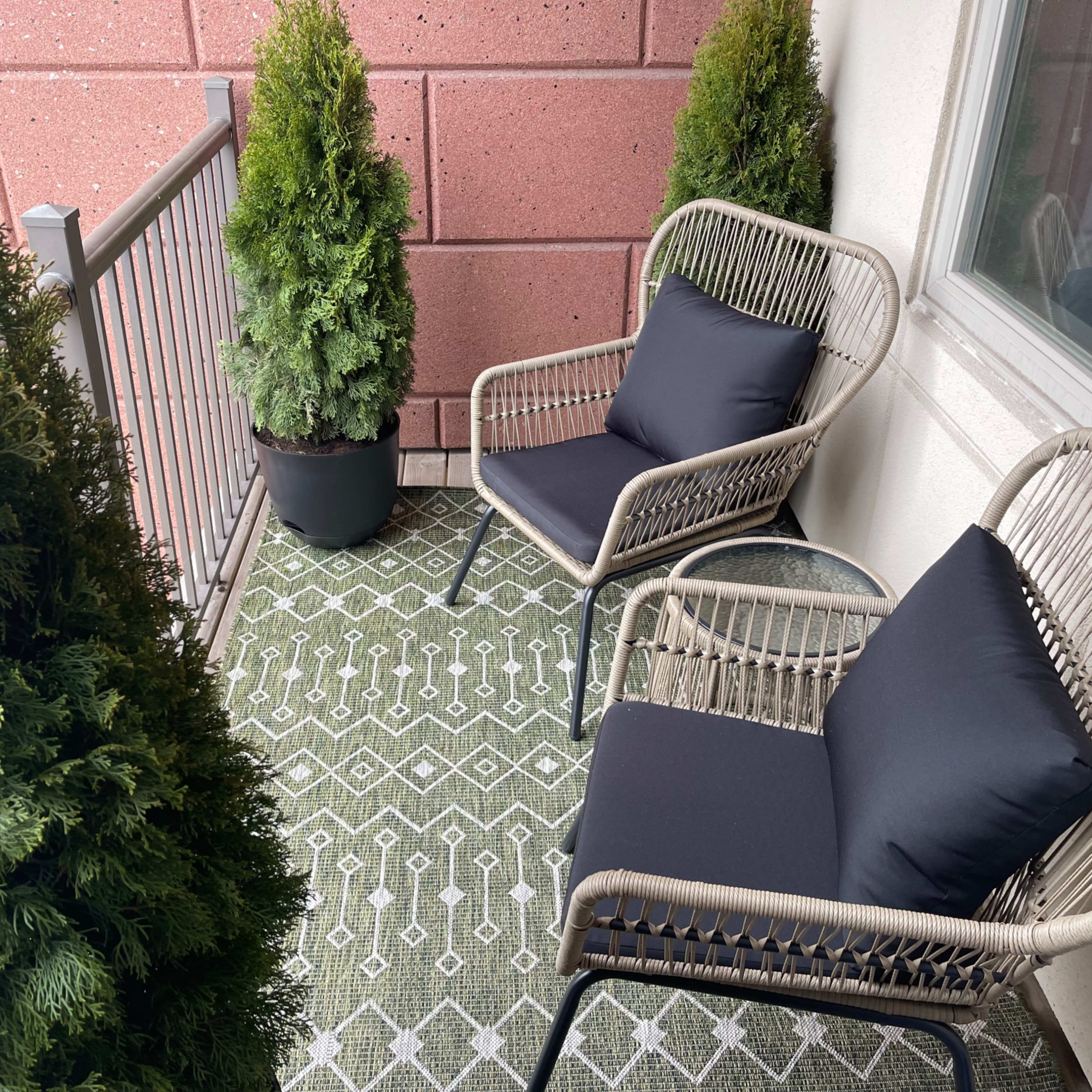 A small balcony featuring two wicker chairs with dark cushions and two potted evergreen plants on either side.