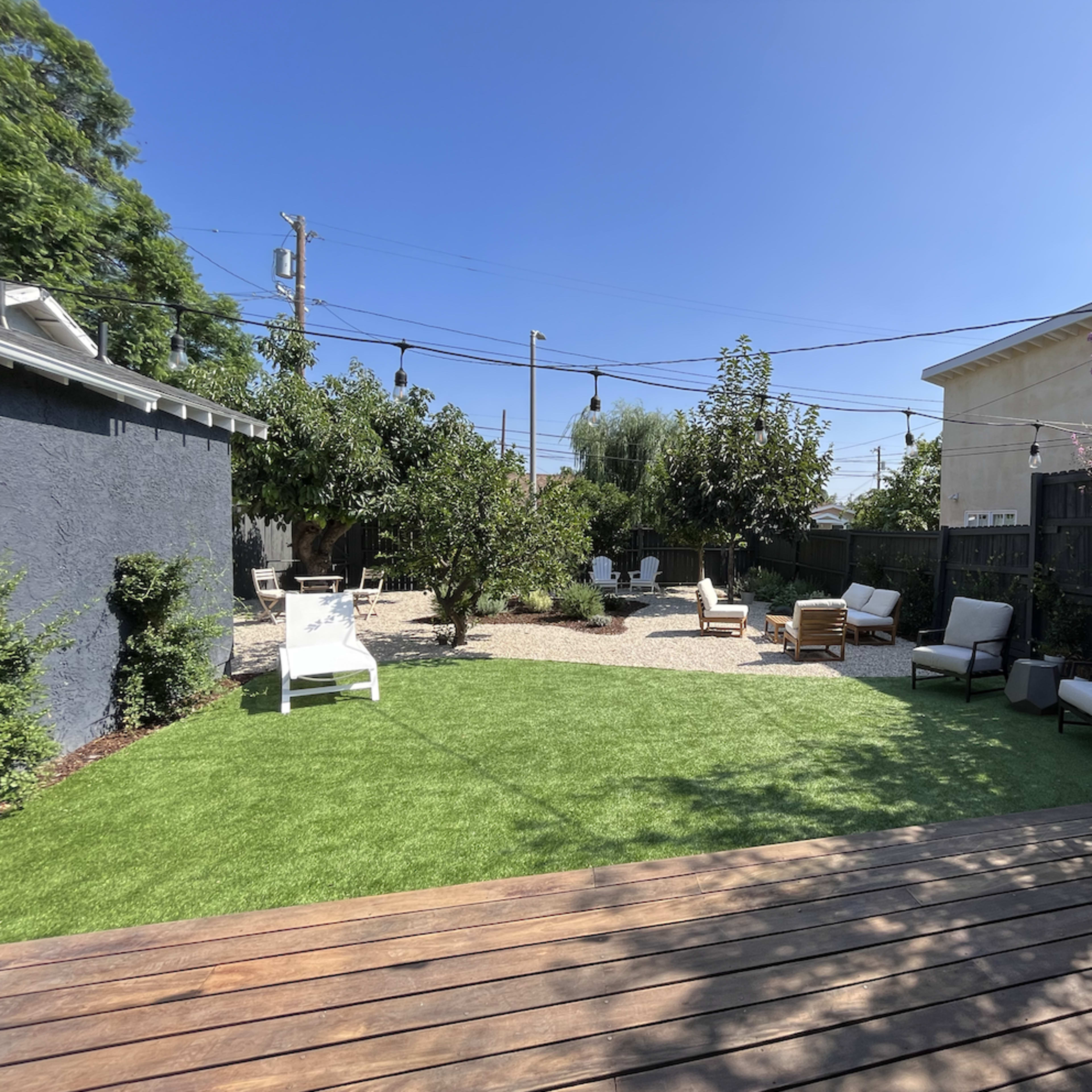 A landscaped outdoor space featuring artificial grass, seating areas with white lounge chairs and wooden furniture, and various trees in a brightly lit setting.