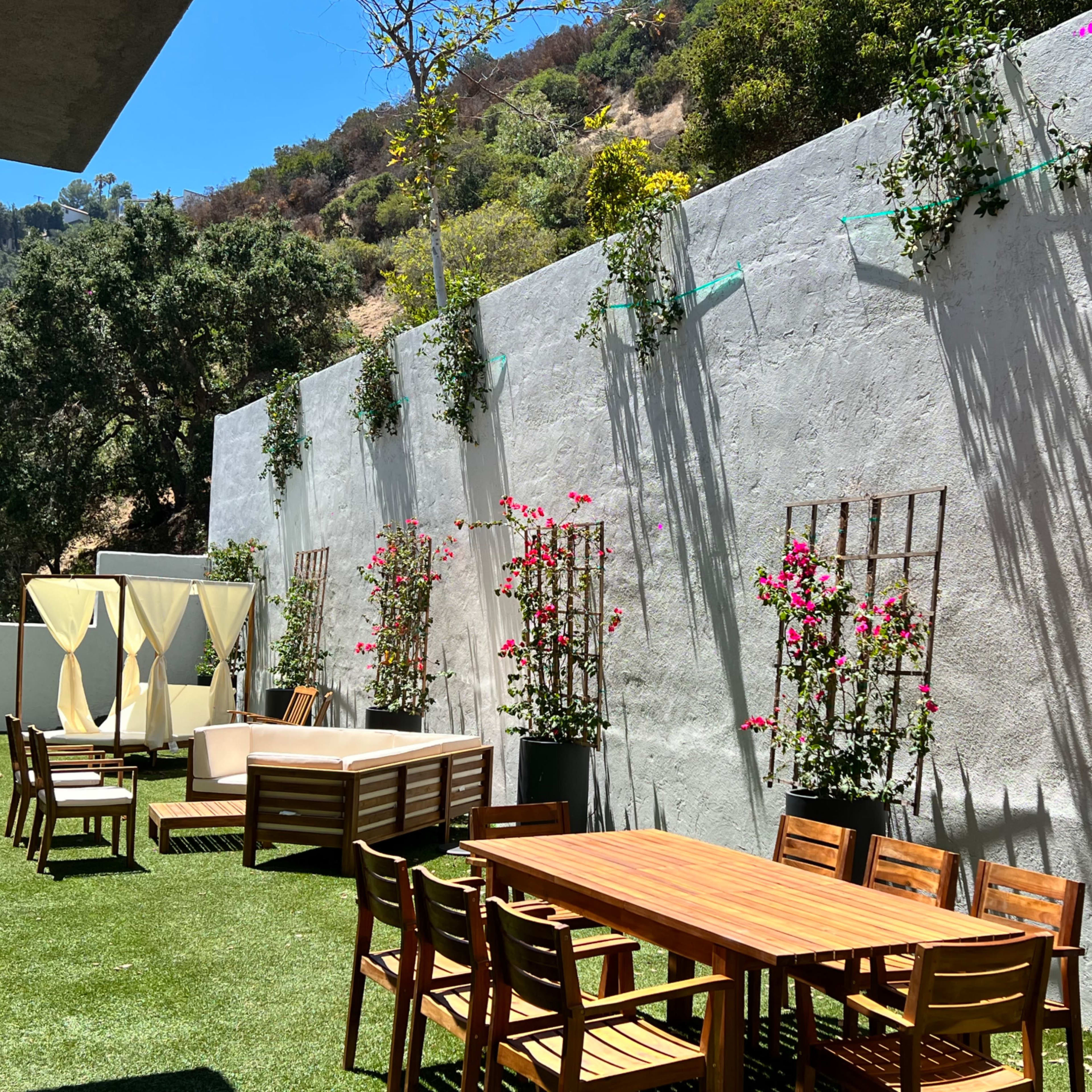 A landscaped outdoor patio featuring a wooden dining table surrounded by chairs, a lounging area with sofas, and vertical planters with pink flowers against a textured wall.