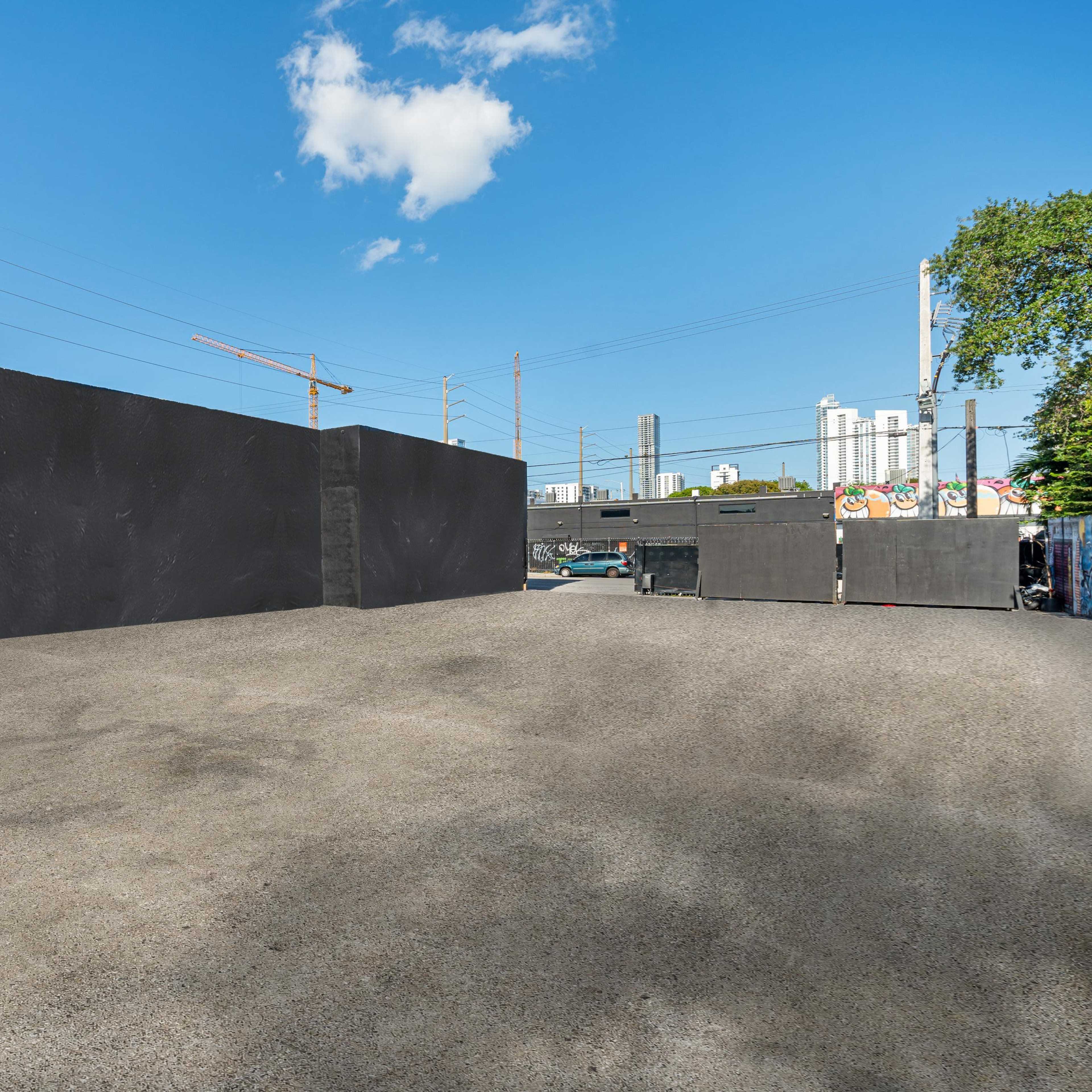 A large, barren concrete area enclosed by high, dark walls, with a city skyline and construction cranes visible in the background.