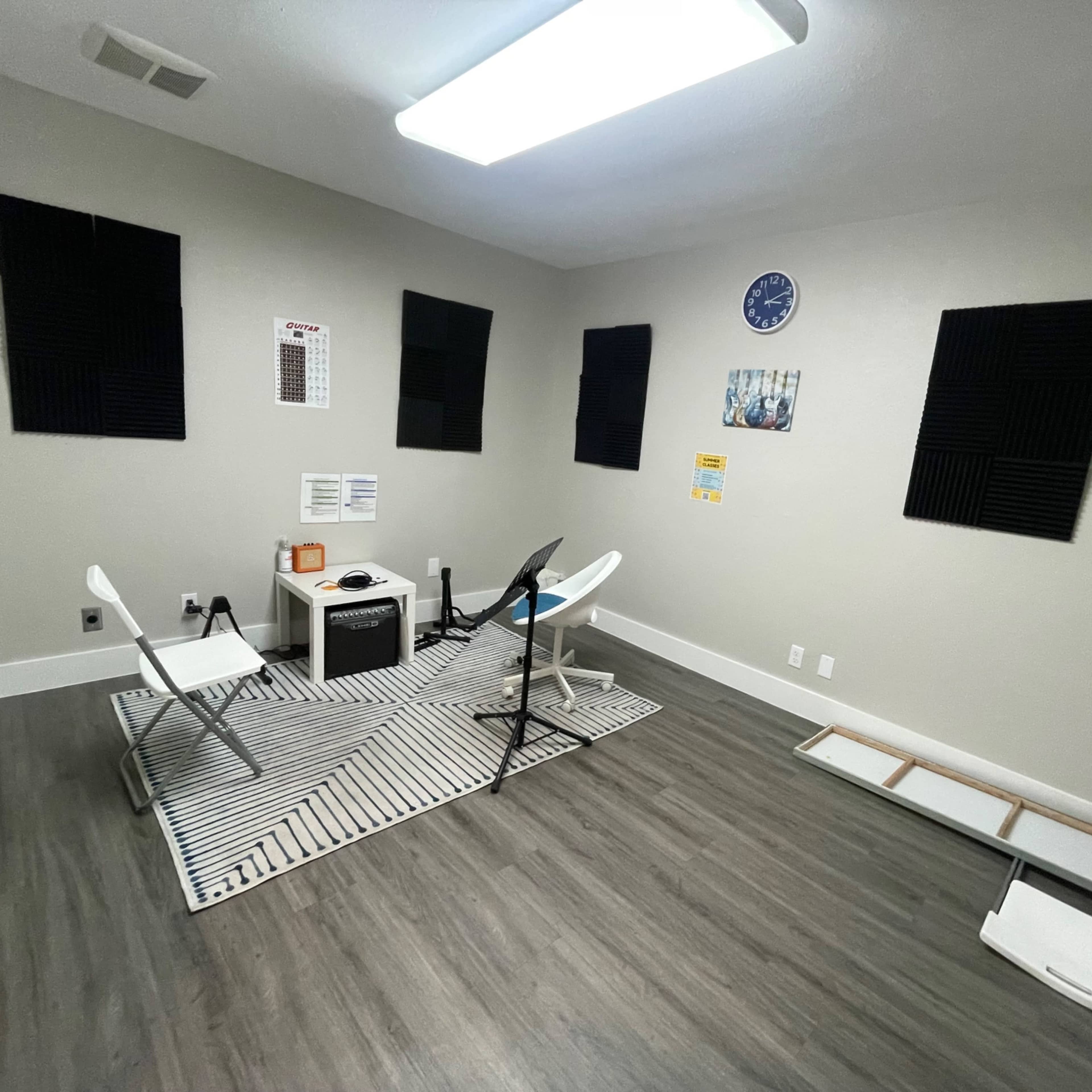 The room features gray walls, a patterned rug, two white chairs, a small table, acoustic foam panels on the walls, and a wall clock.