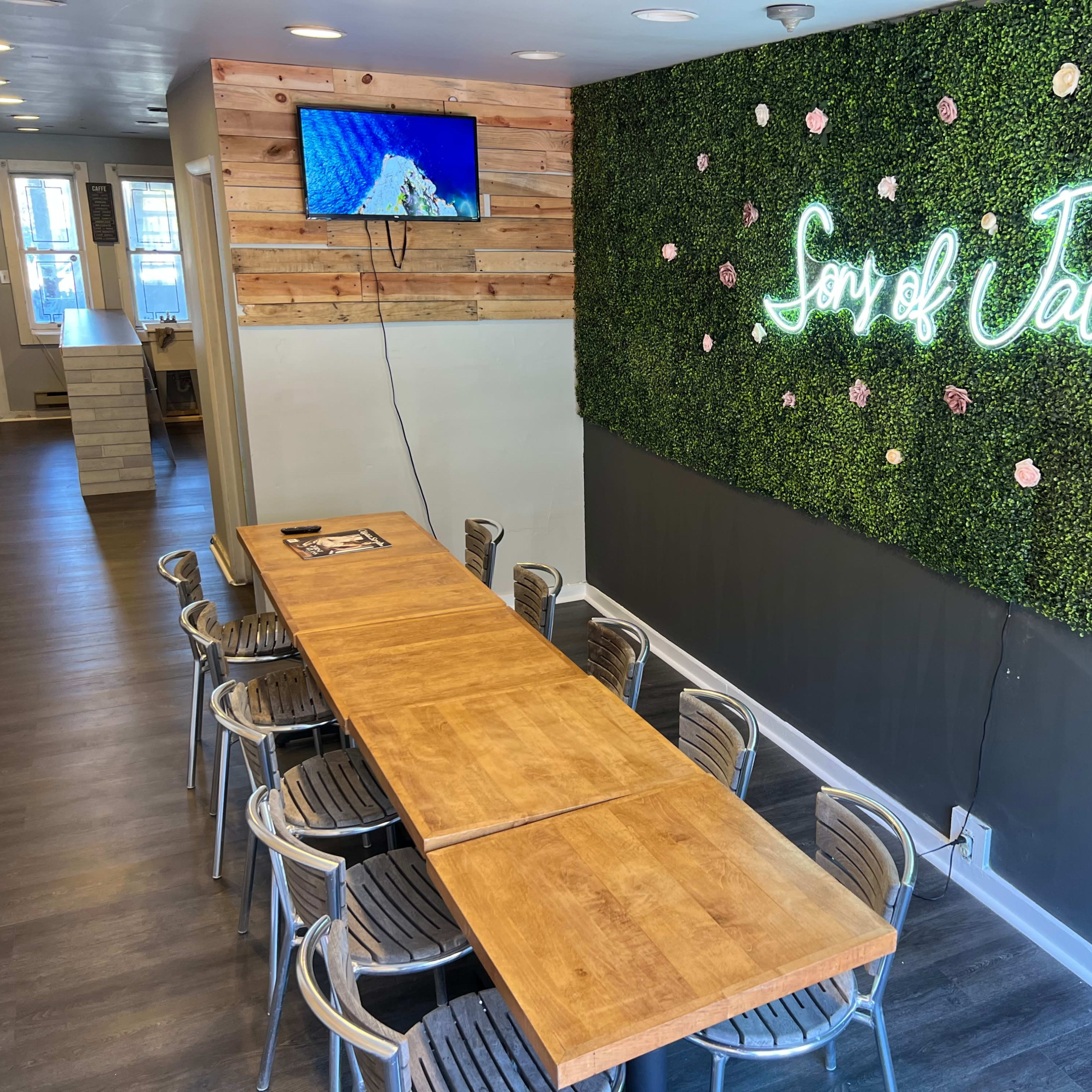 A long wooden table with metal chairs is set against a green wall adorned with artificial foliage and illuminated signage reading "Son of Java."