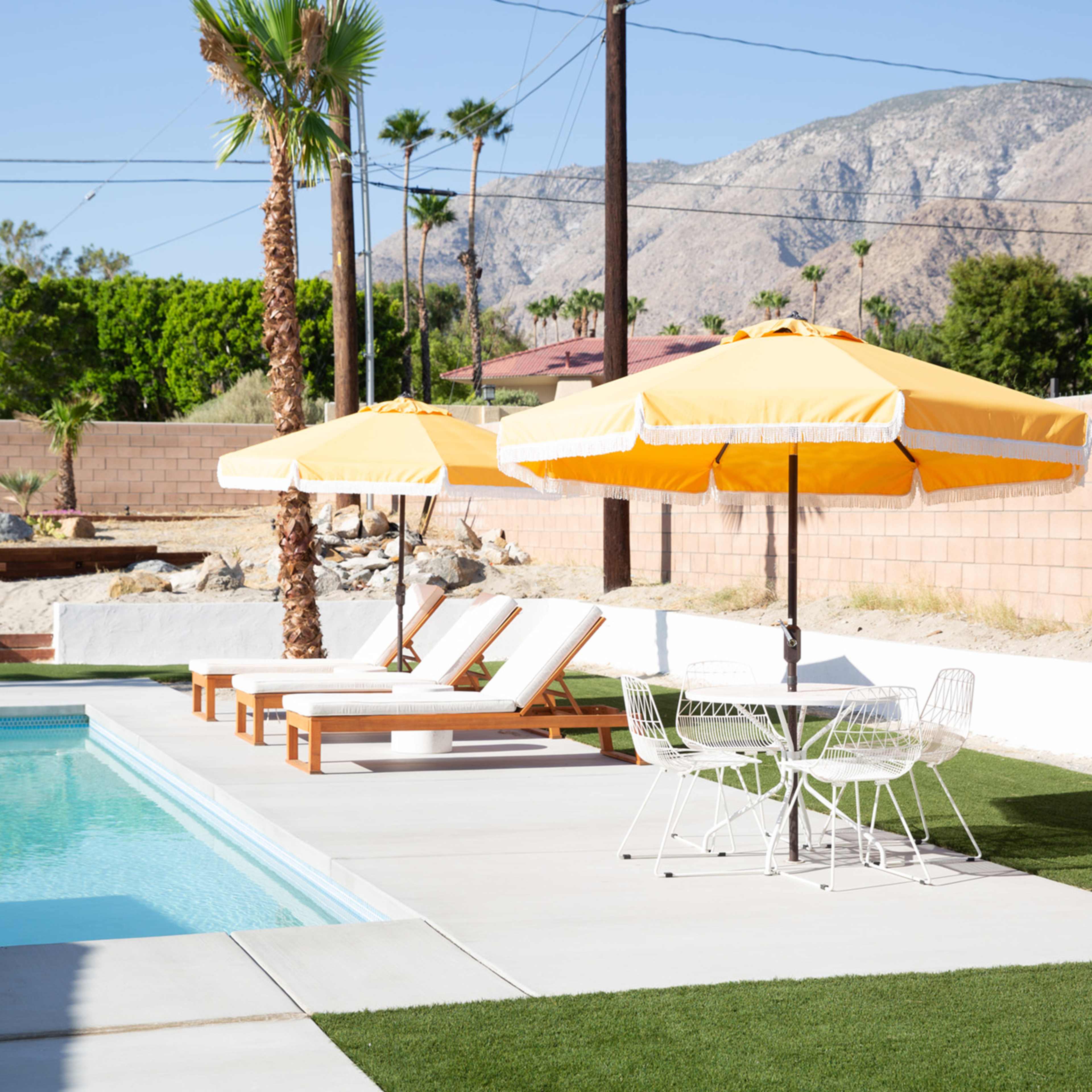 A swimming pool with two yellow umbrellas and lounge chairs nearby, surrounded by landscaped greenery and mountains in the background.