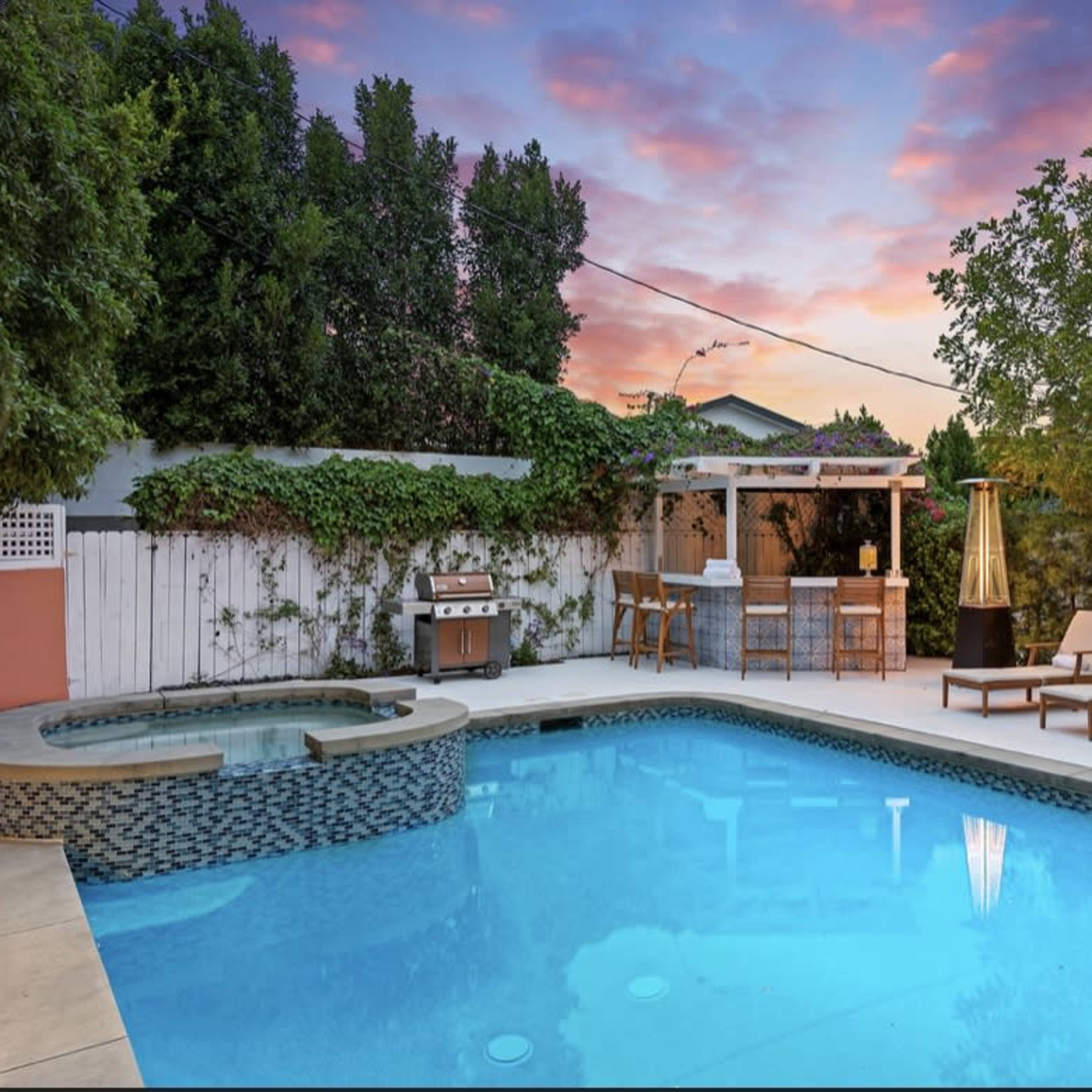 A backyard pool area with a spa, lounge chairs, a grill, and a shelter, set against a backdrop of a colorful sunset.